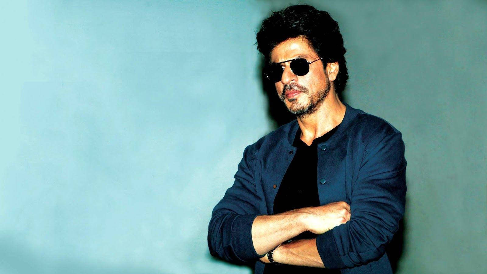Shahrukh 桌布 壁紙 — 一名深色頭髮、戴著太陽眼鏡的男子，自信地交叉雙臂，站在純藍色背景前。