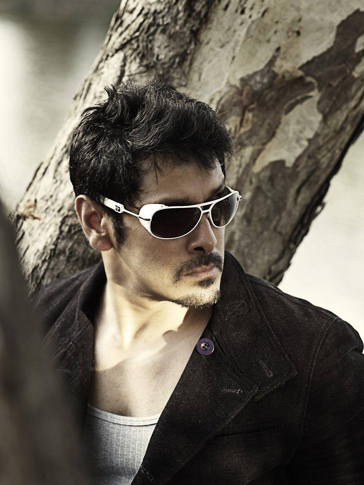 Sunglasses 桌布 壁紙 — 一名深色頭髮、戴著太陽眼鏡的男子，在柔和自然光線下靠著紋理分明的樹幹。