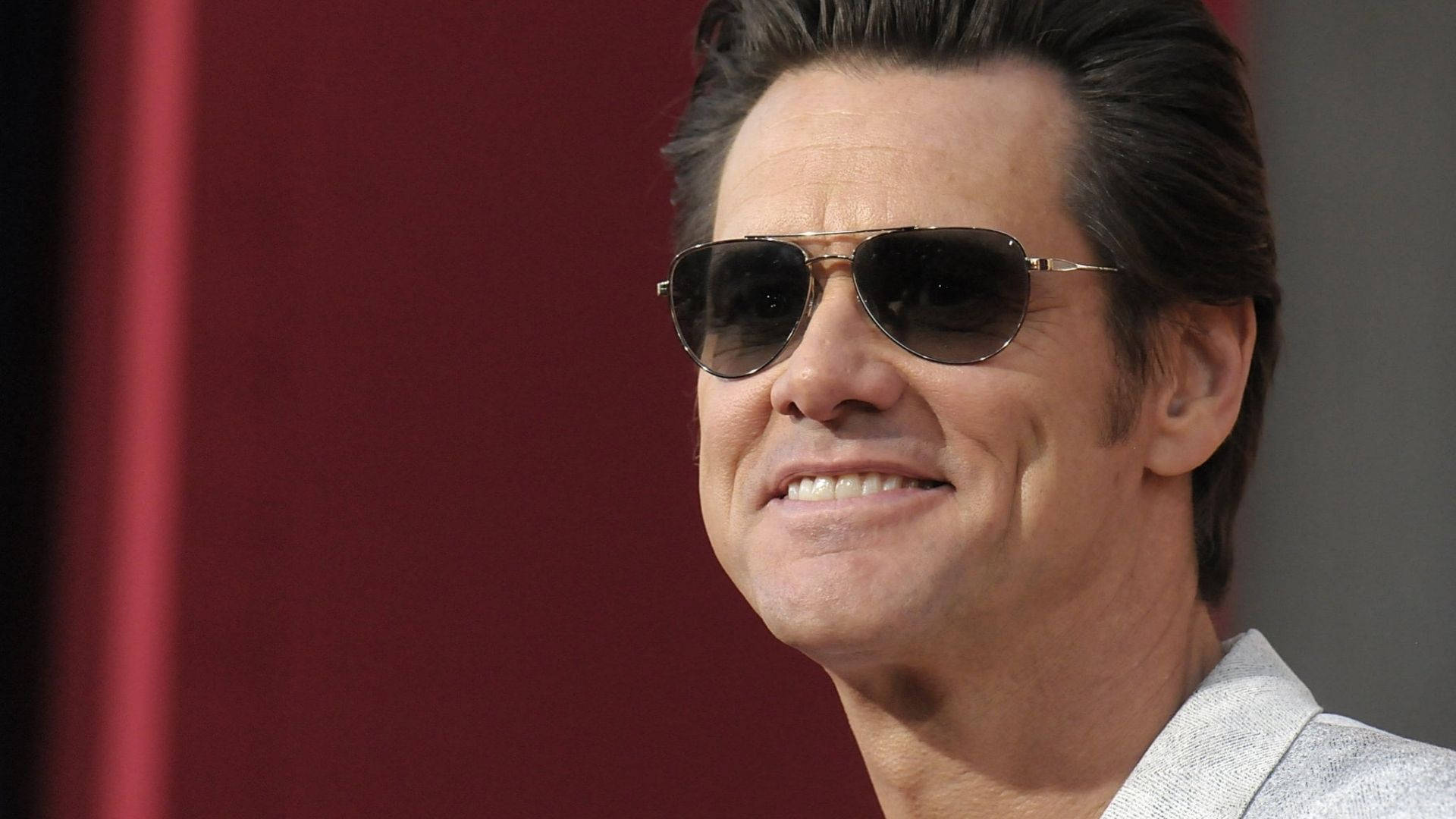 Jim Carrey 桌布 壁紙 — 一名深色頭髮、戴著飛行員太陽眼鏡的男子在深紅色背景前溫暖地微笑。