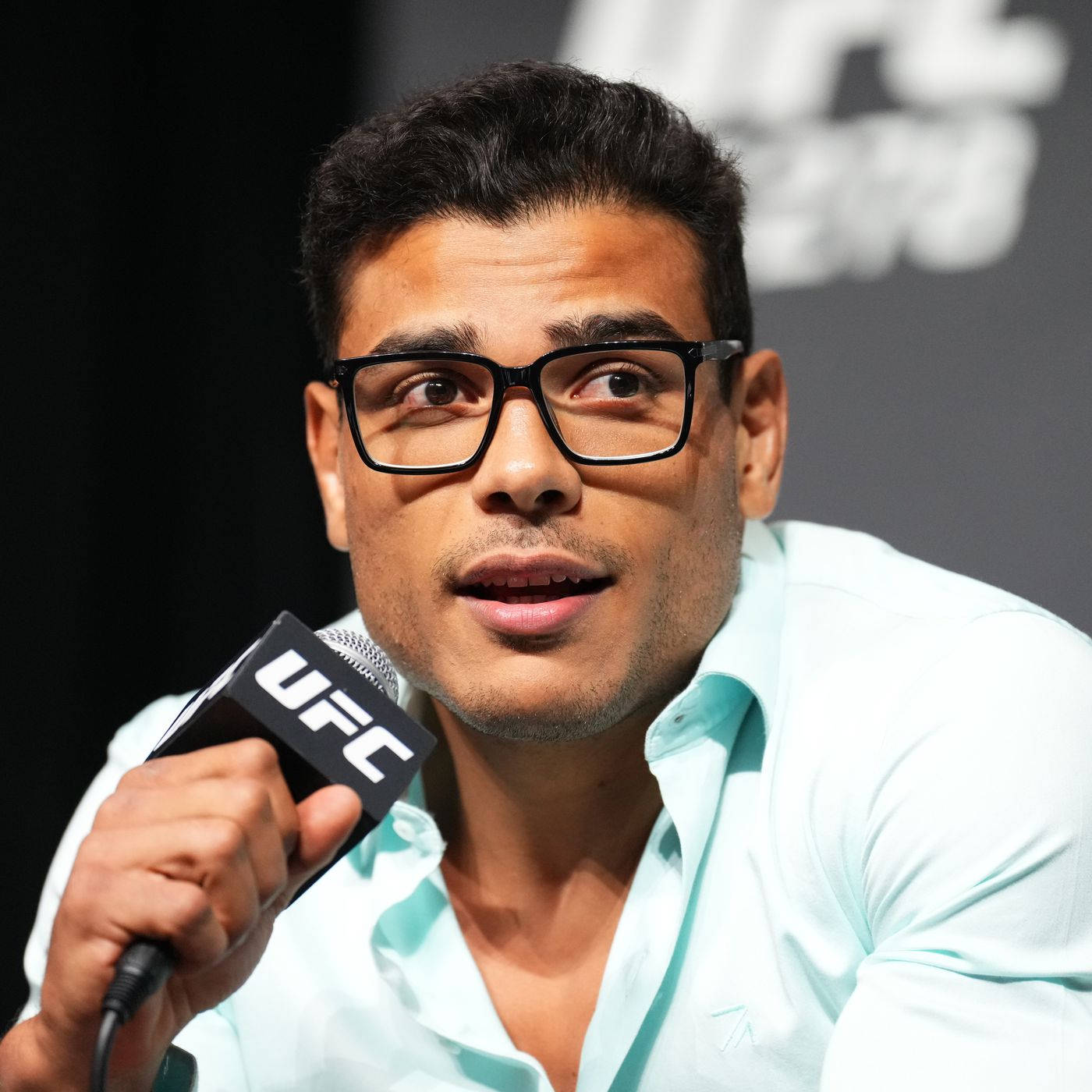 UFC 桌布 壁紙 — 一名深色頭髮、戴黑色眼鏡的男子，身穿淺藍色襯衫，在記者會上對著UFC品牌麥克風發言。