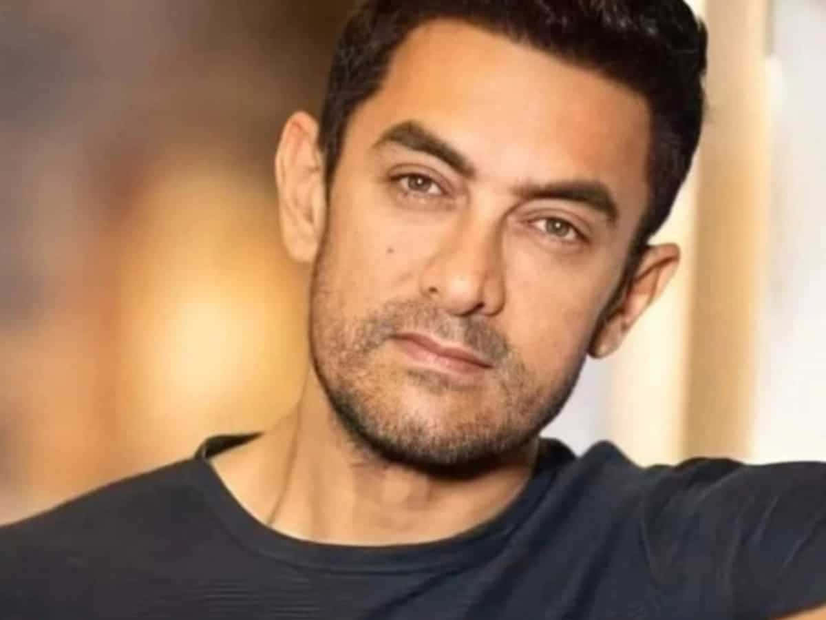 Aamir Khan 桌布 壁紙 — 一名深色頭髮、留著鬍渣的男子穿著黑色T恤的特寫肖像，背景是柔和模糊的暖色調。