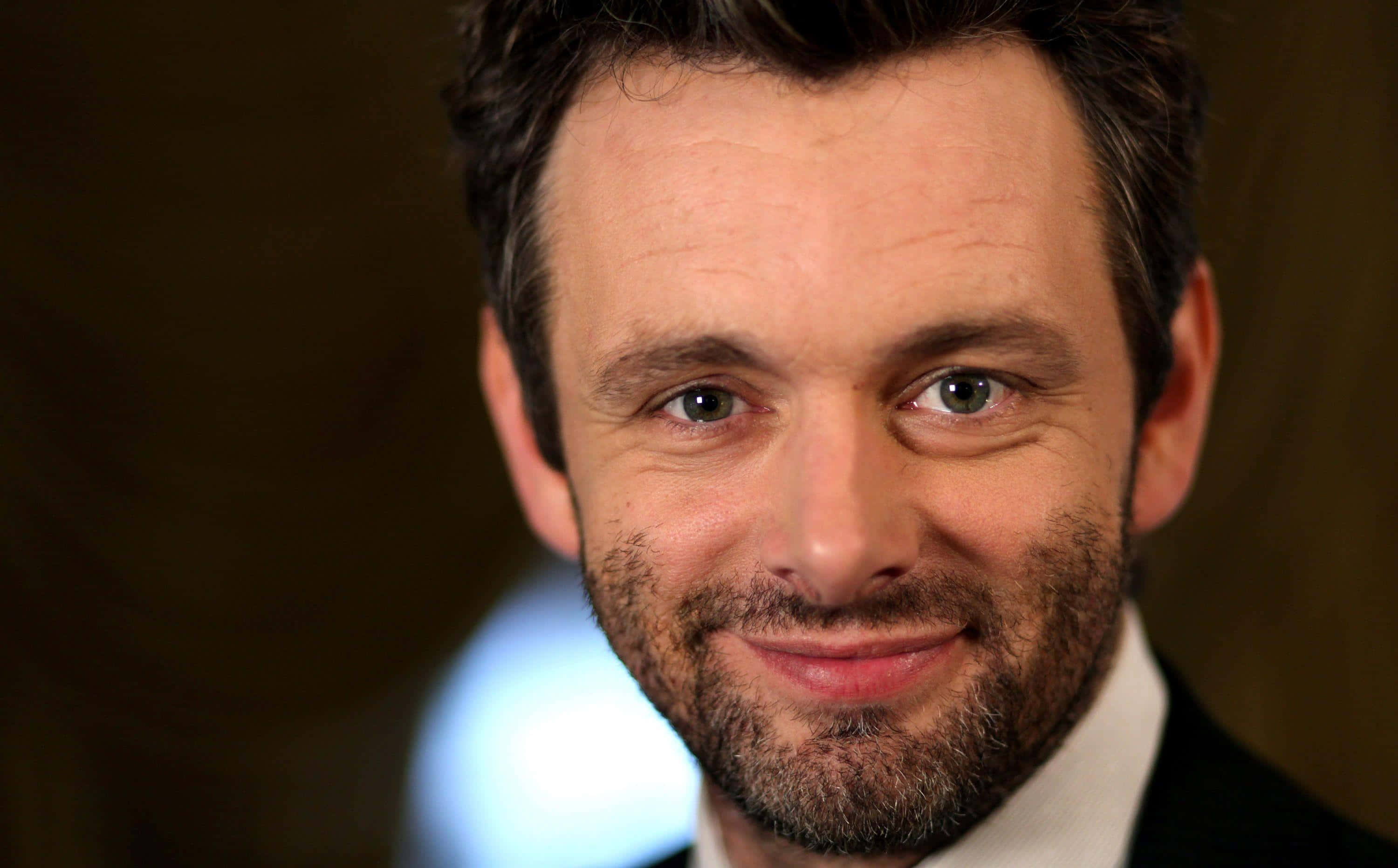 Michael Sheen 桌布 壁紙 — 一名深色頭髮、留鬍鬚、面帶微笑的男士穿著西裝，在模糊的溫暖背景前拍攝的特寫肖像。
