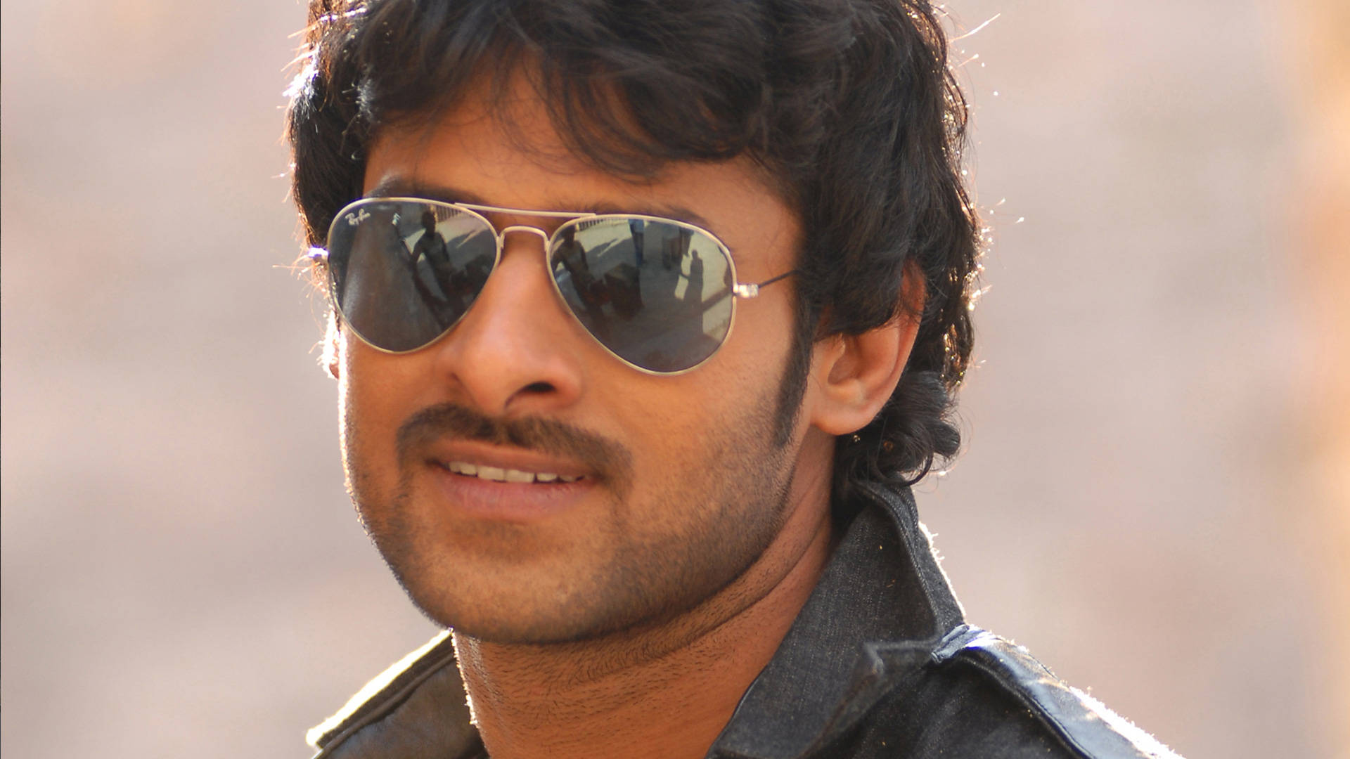 Prabhas HD 桌布 壁紙 — 一名深色頭髮、留鬍鬚的男子，在模糊的暖色背景前戴著飛行員太陽眼鏡和深色外套。