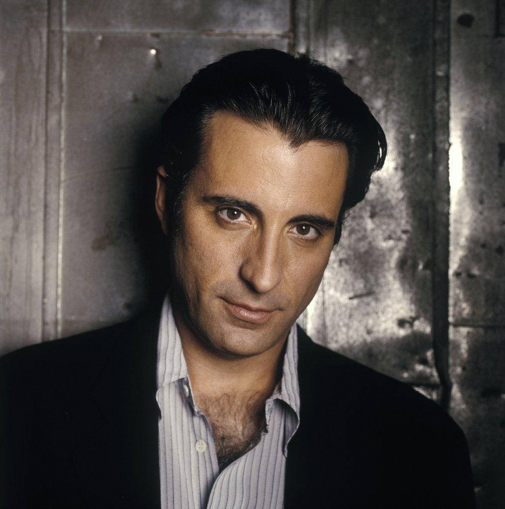Andy Garcia 桌布 壁紙 — 一名深色頭髮、眼神銳利的男子，在充滿氛圍的工作室肖像中靠著紋理金屬牆。