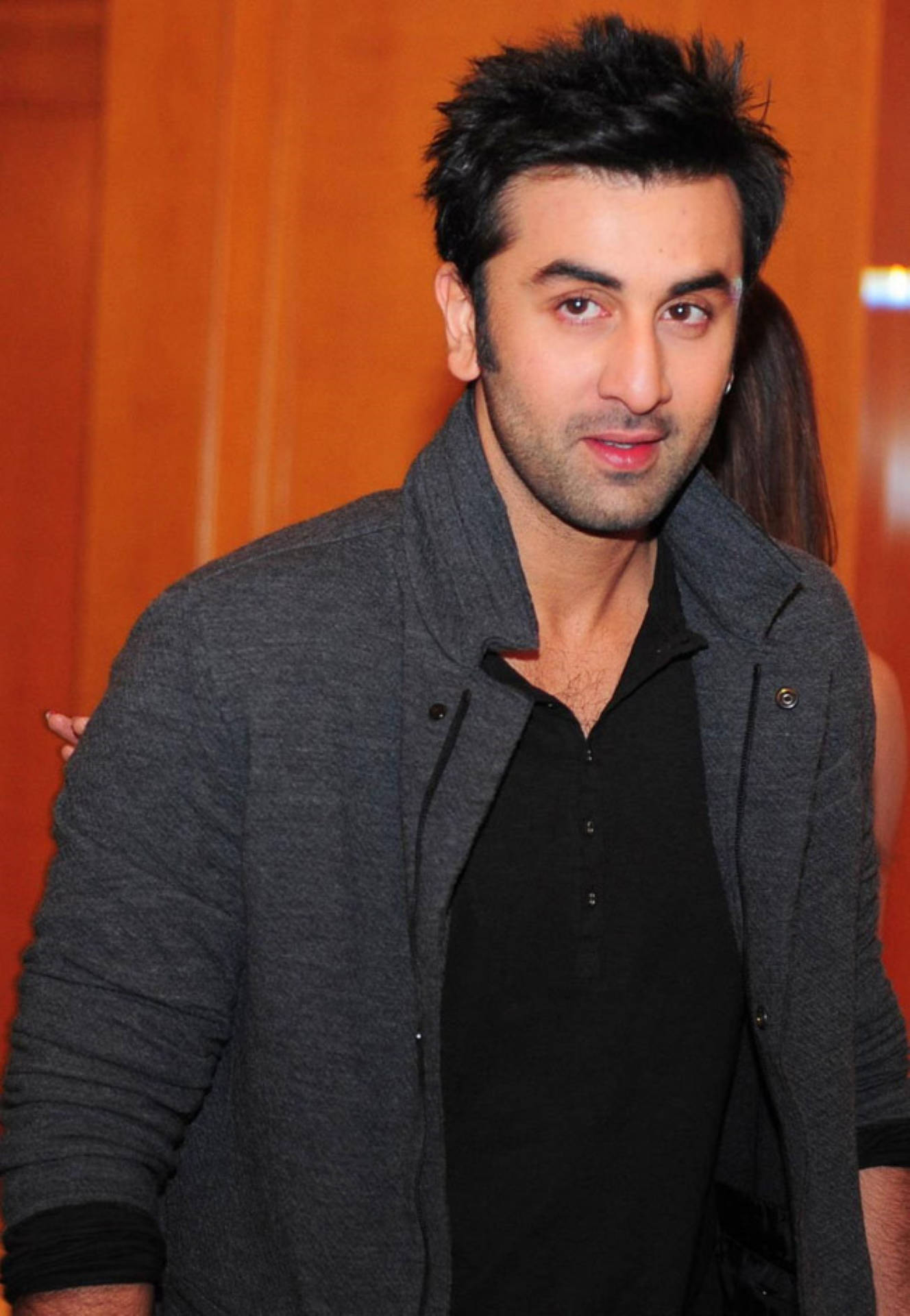Ranbir Kapoor 桌布 壁紙 — 一名深色頭髮、鬍渣的男人，在溫暖的橙色背景前，穿著灰色外套內搭黑色襯衫。