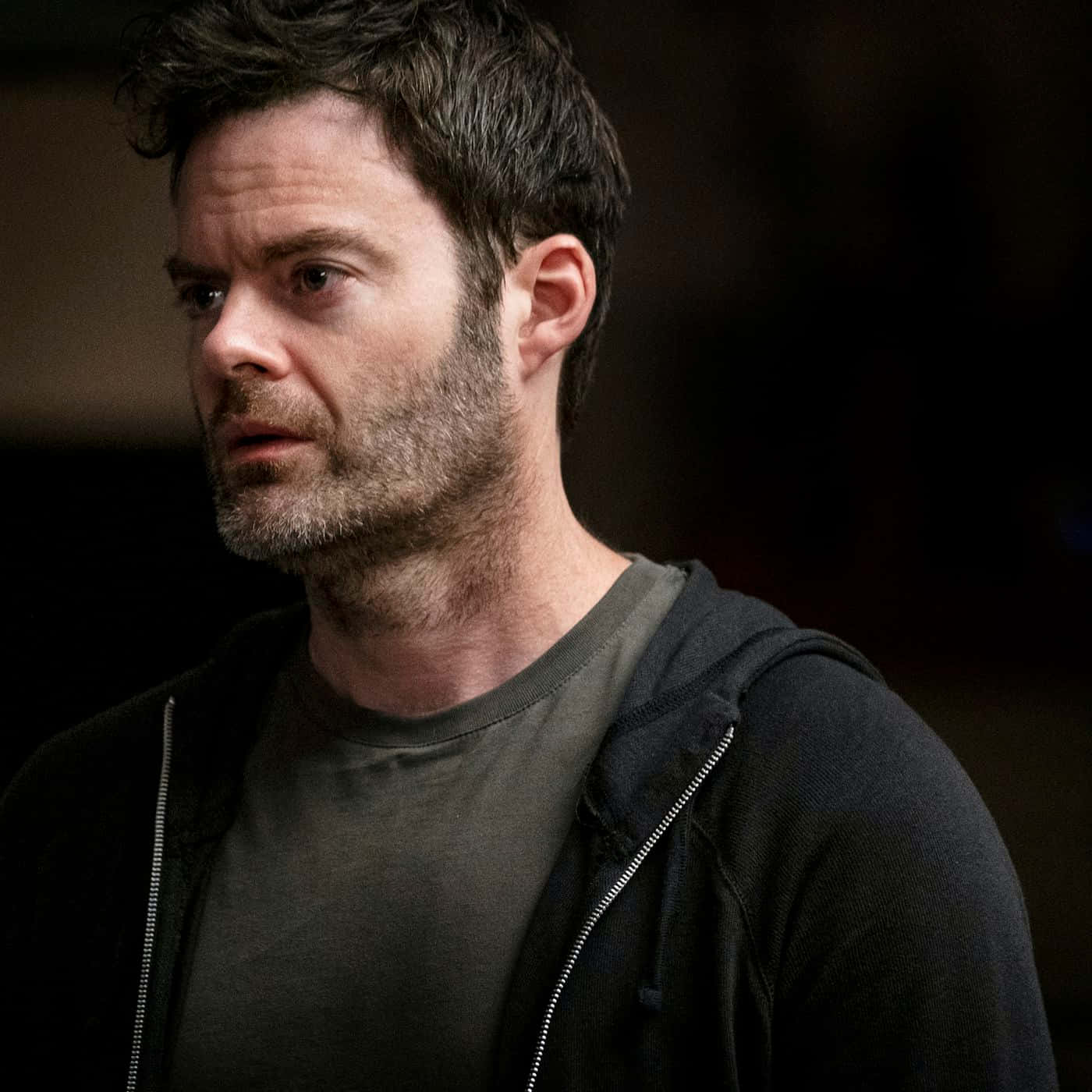 bill hader 桌布 壁紙 — 一名深色頭髮、鬍渣的男子穿著黑色連帽衫，內搭灰色襯衫，在昏暗的燈光下以嚴肅的表情看向鏡頭外。