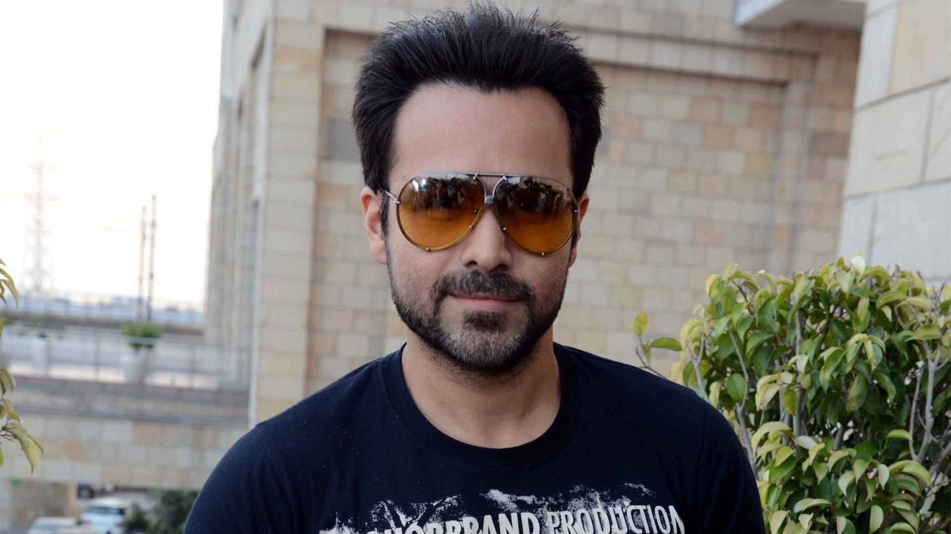 Emraan Hashmi 桌布 壁紙 — 一名深色頭髮和鬍鬚的男子，在戶外靠近綠色灌木和石建築物的地方，戴著飛行員太陽眼鏡和黑色T恤。