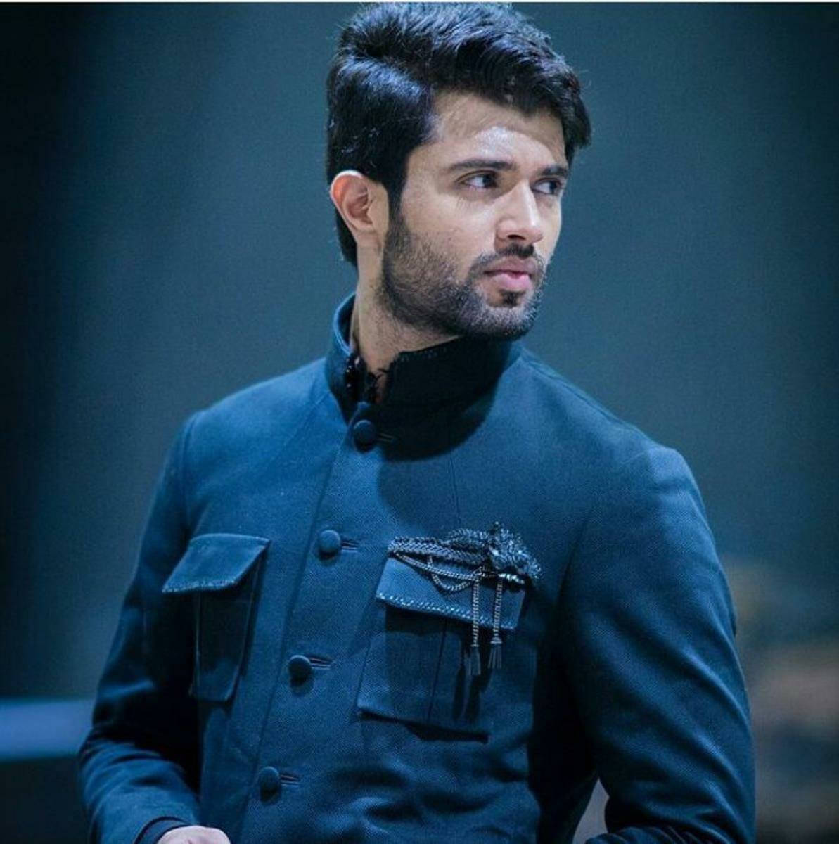 vijay devarakonda 4K 桌布 壁紙 — 一名深色頭髮和鬍鬚的男子，穿著時尚的深藍色傳統夾克，帶有裝飾性鏈條點綴，背景柔和。