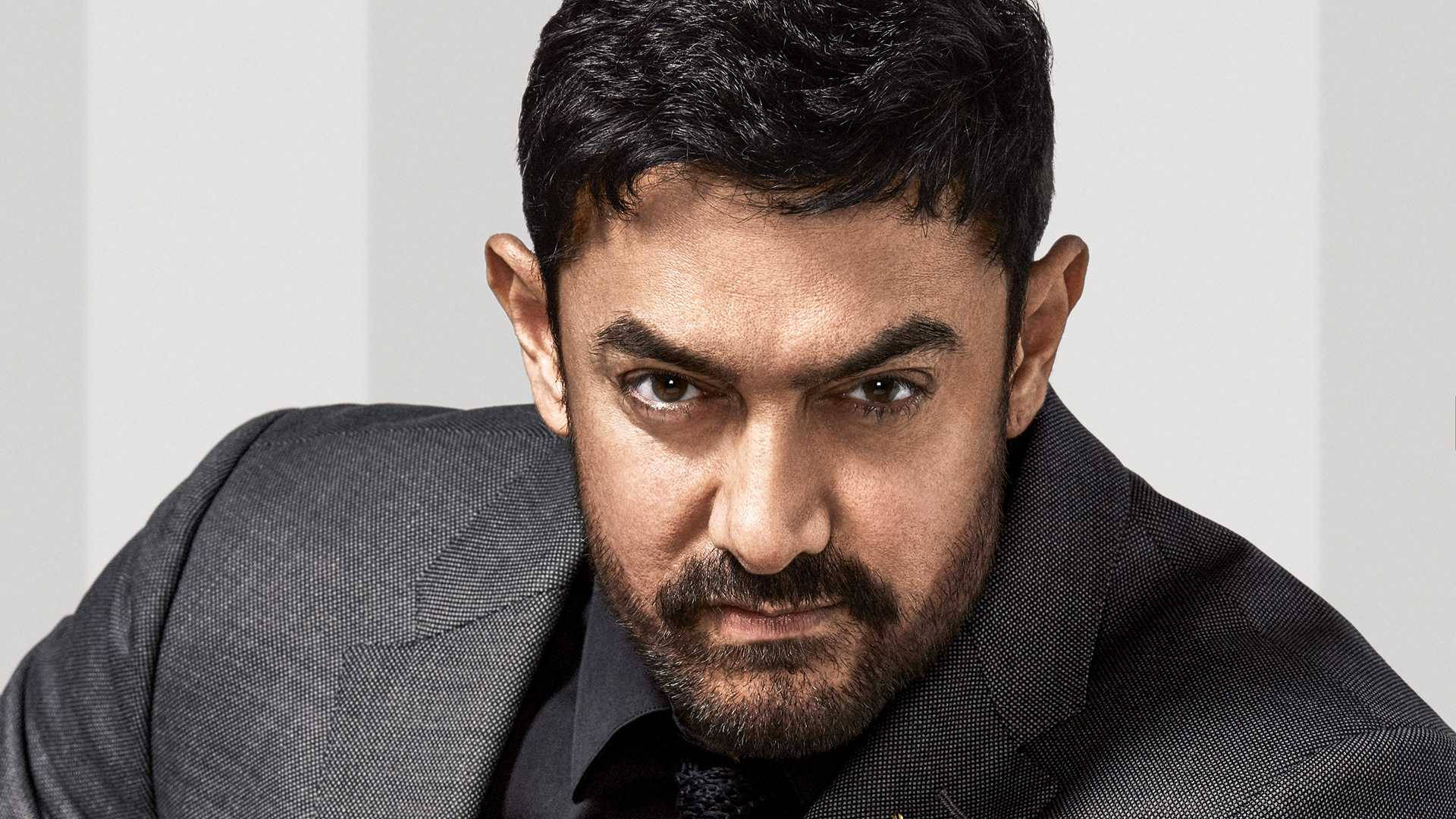 Aamir Khan 桌布 壁紙 — 一名深色頭髮和鬍鬚的男子，穿著深色西裝，在純色淺色背景前，凝視著鏡頭。