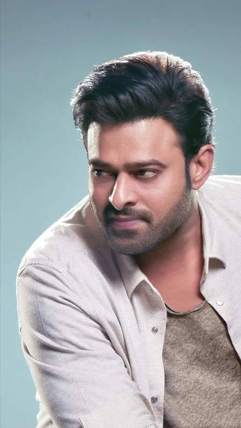 Prabhas 桌布 壁紙 — 一名深色頭髮和鬍鬚的男子，穿著淺色襯衫，在柔和的藍色背景前，表情嚴肅。