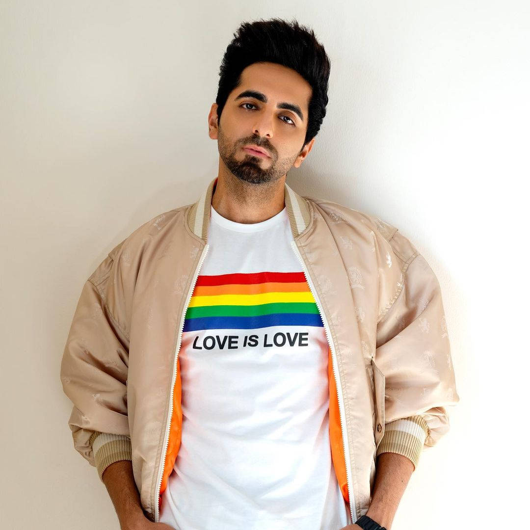 ayushmann khurrana 桌布 壁紙 — 一名深色頭髮和鬍鬚的男子，身穿米色飛行員夾克，內搭白色T恤，上面有彩虹條紋和「love is love」字樣，背景是純白牆壁。