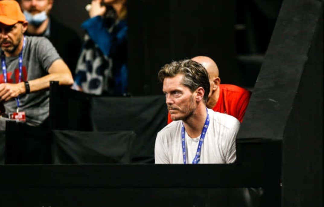Tennis 桌布 壁紙 — 一名深色頭髮和鬍鬚的男子在昏暗的競技場中專注地坐著，掛著掛繩，周圍有其他人在觀看。