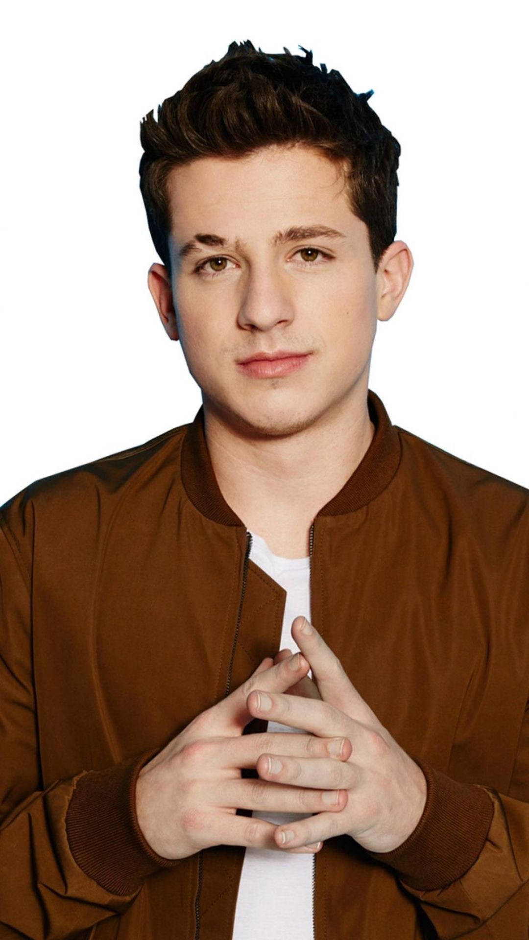 Charlie Puth 桌布 壁紙 — 一名深色頭髮的年輕男子身穿棕色飛行員夾克，內搭白色T恤，雙手合十放在身前，背景為純白色。