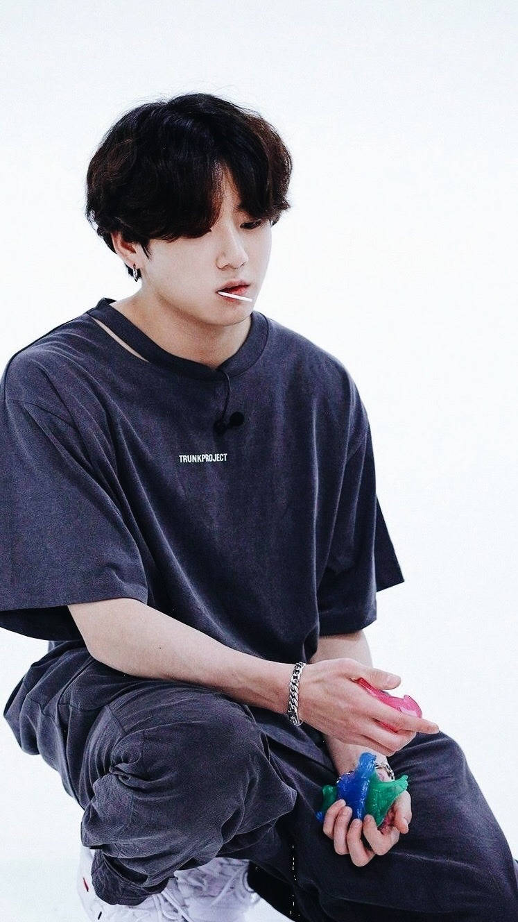 Jungkook 桌布 壁紙 — 一名深色頭髮的年輕男子隨意地坐在純白色背景前，手持色彩繽紛的物件。