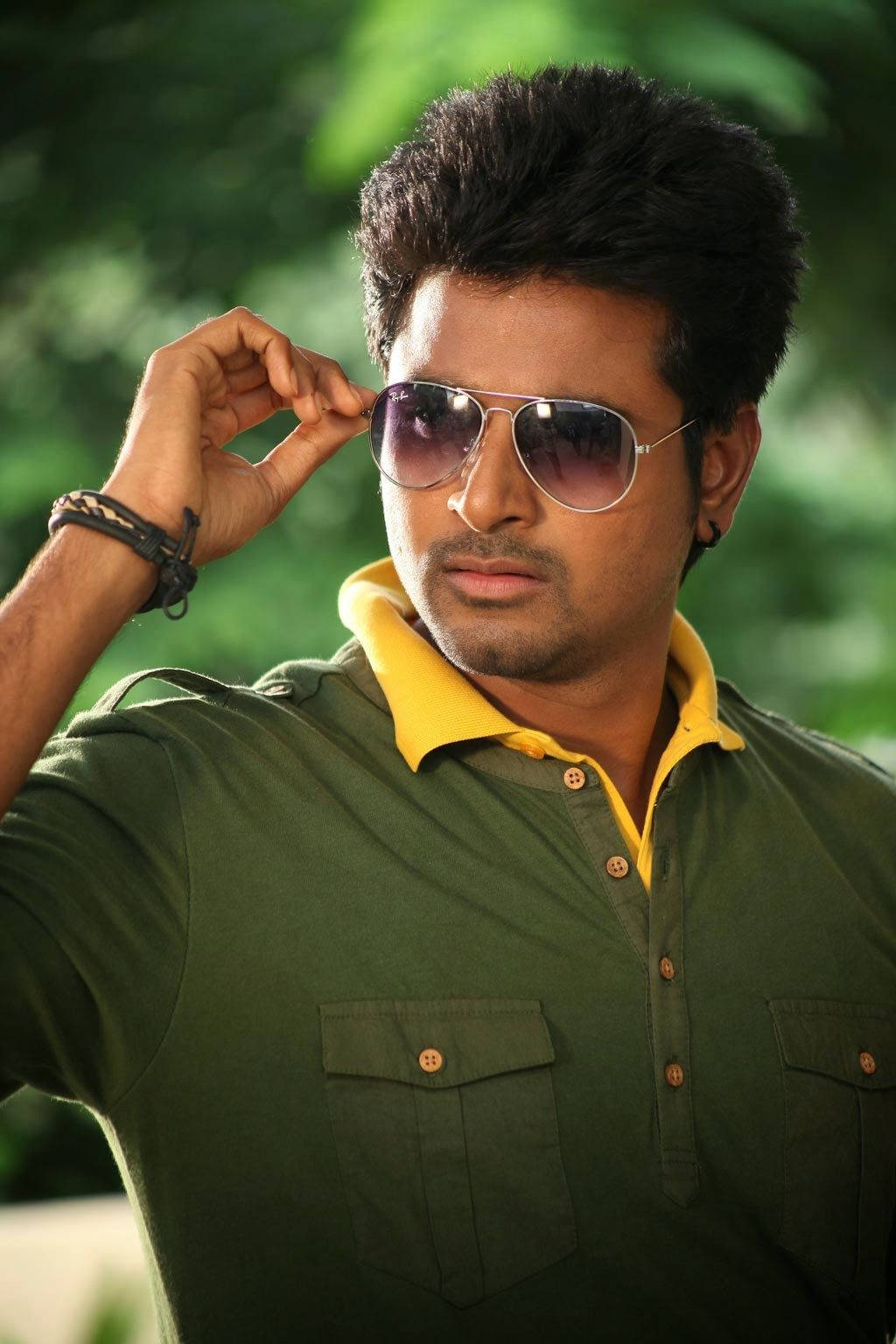 Sivakarthikeyan 桌布 壁紙 — 一名深色頭髮的男子在模糊的綠色樹葉背景前，穿著帶有黃色領子的綠色Polo衫，調整他的飛行員太陽眼鏡。