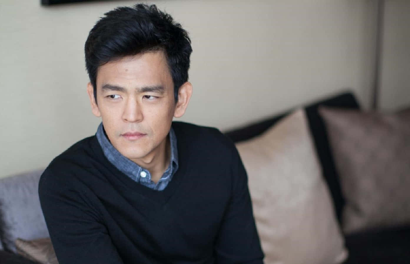 John Cho 桌布 壁紙 — 一名深色頭髮的男子坐在沙發上，身穿黑色毛衣內搭藍色襯衫，若有所思地望向一旁。