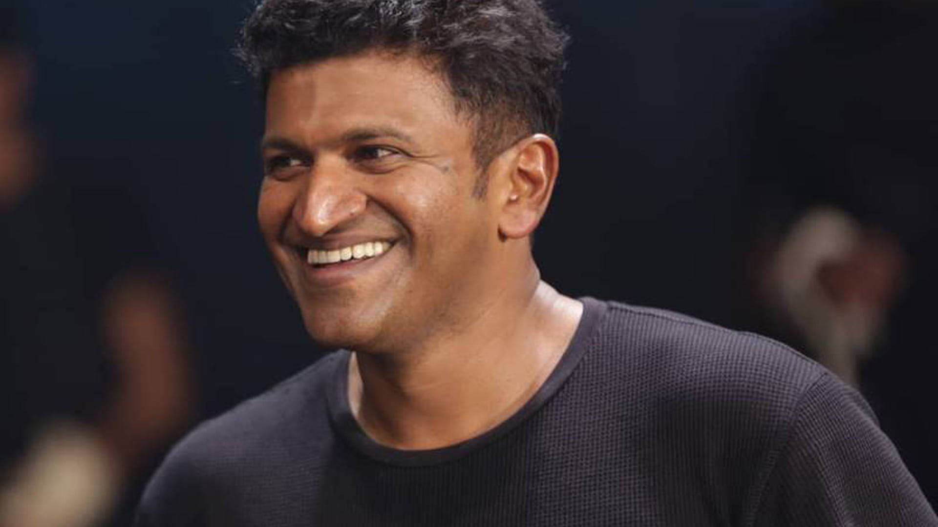 Puneeth Rajkumar 桌布 壁紙 — 一名深色頭髮的男子穿著深色質感襯衫，在模糊的背景前溫暖地微笑。