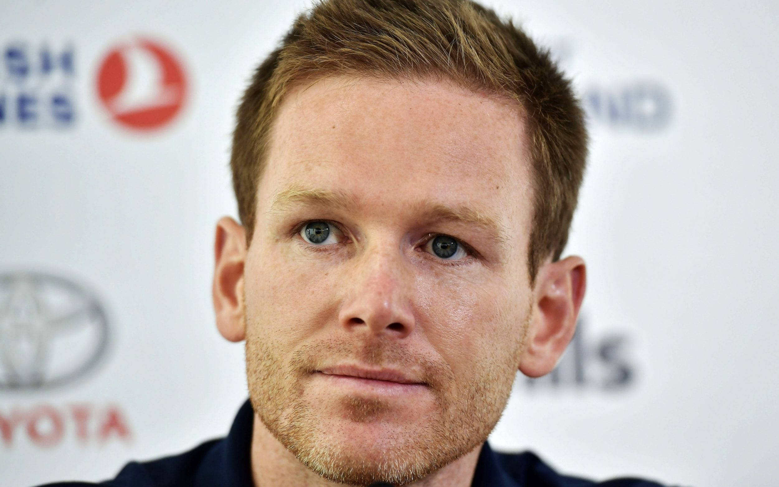 eoin morgan 桌布 壁紙 — 一名淺棕色頭髮、藍眼睛的男子在品牌背景前直視鏡頭的近距離肖像。