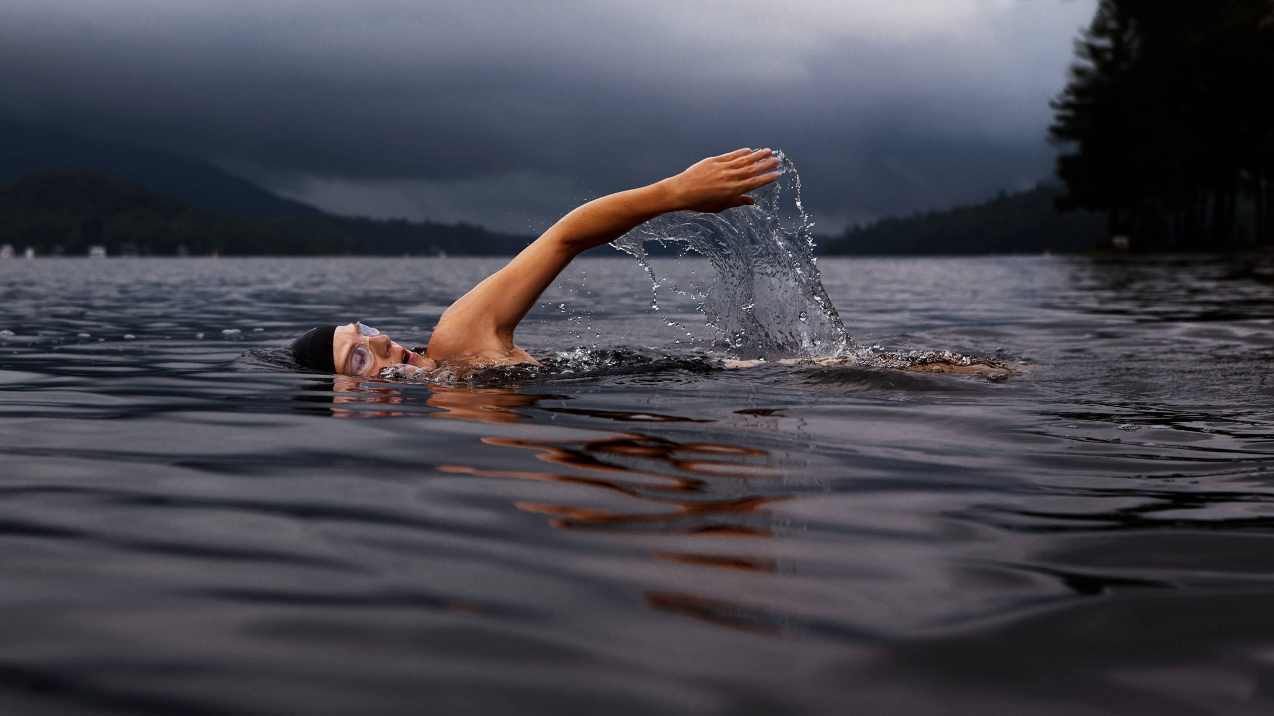 Swimming 桌布 壁紙 — 一名游泳者在風暴天空下的黑暗水中滑行，背景有山巒若隱若現。