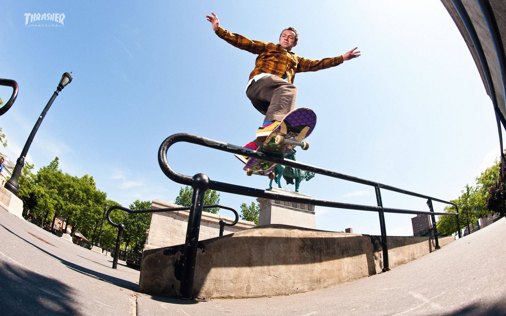 Thrasher 桌布 壁紙 — 一名滑板手在半空中，在黑色欄杆上表演技巧，背景是晴朗的藍天，帶有魚眼鏡頭變形效果。