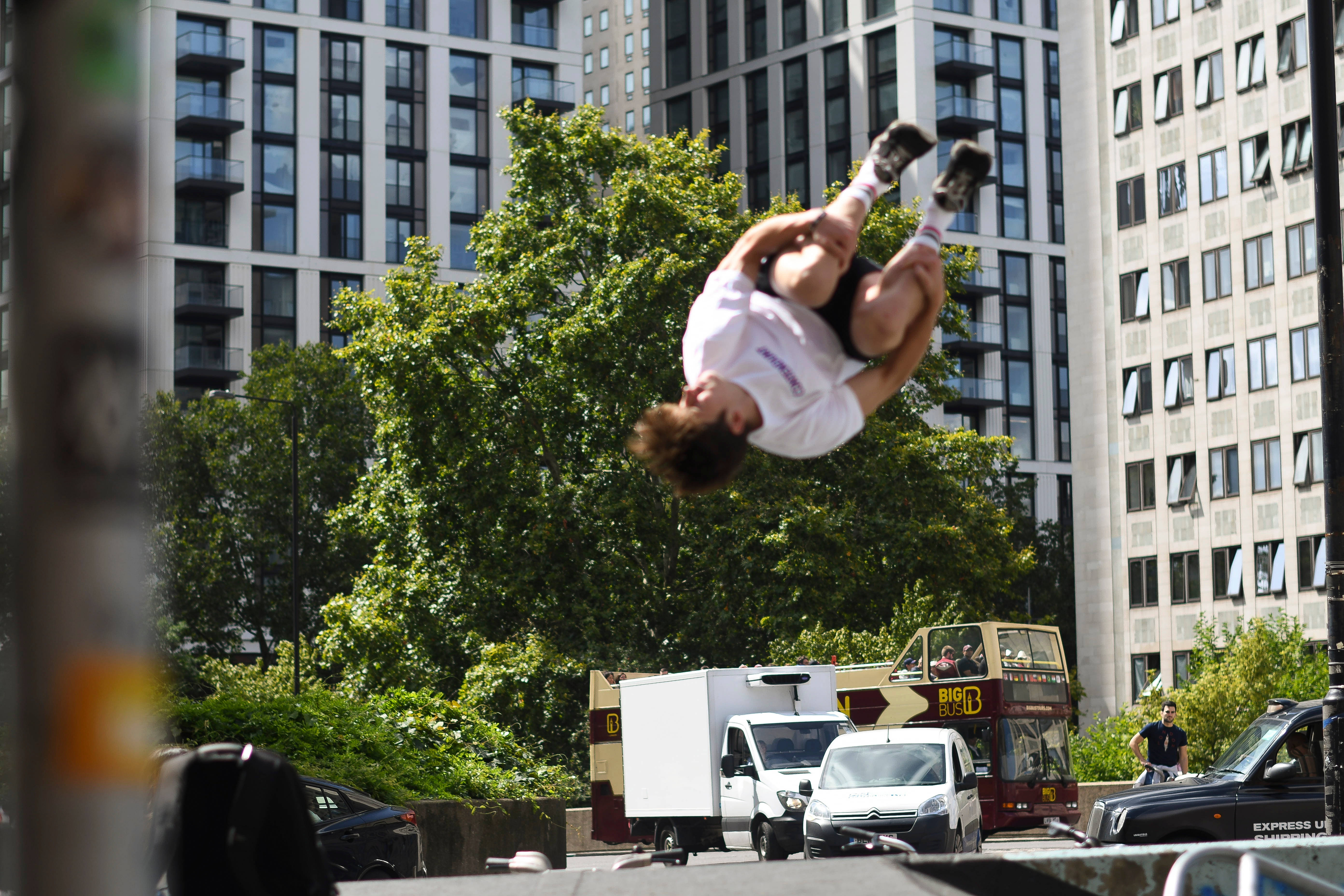 Parkour 桌布 壁紙 — 一名滑板手在半空中於陽光明媚的城市街道上表演後空翻，背景是高樓大廈和綠樹。