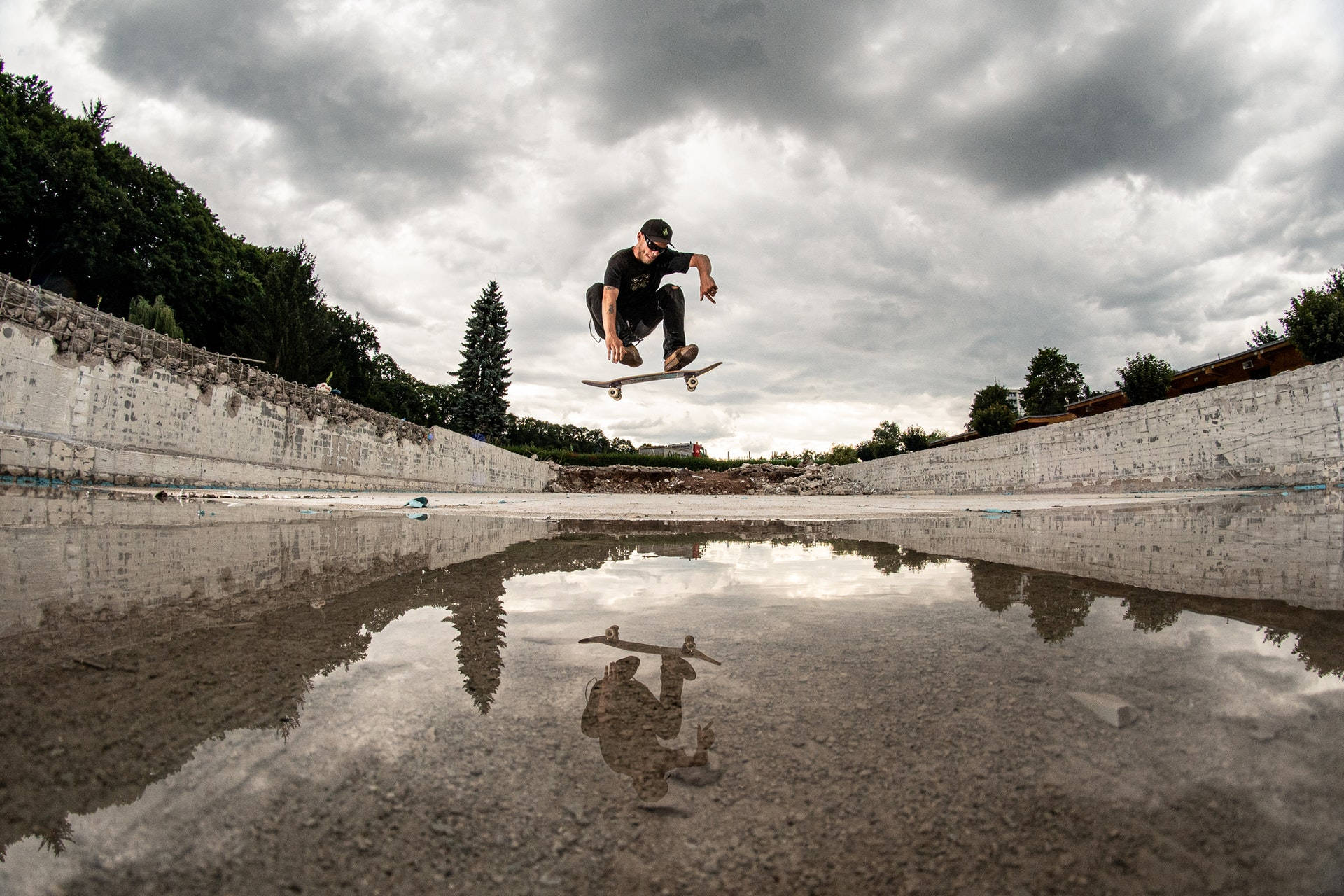 Skater Boy 桌布 壁紙 — 一名滑板手在廢棄混凝土滑板場的水坑上方騰空，天空多雲。