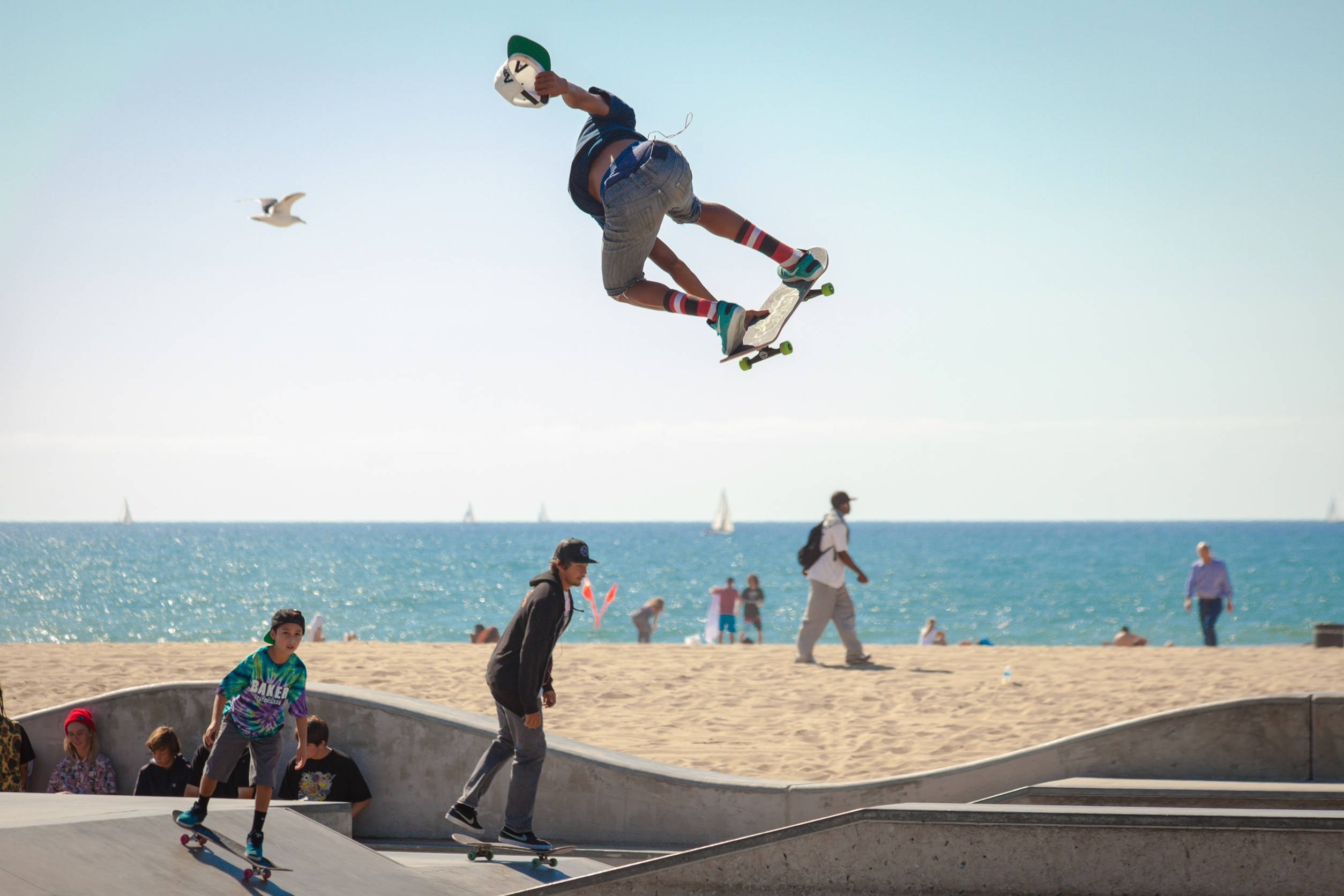 Skater Boy 桌布 壁紙 — 一名滑板手在陽光明媚的海灘上，於混凝土坡道上騰空飛起，背景是海洋和帆船。