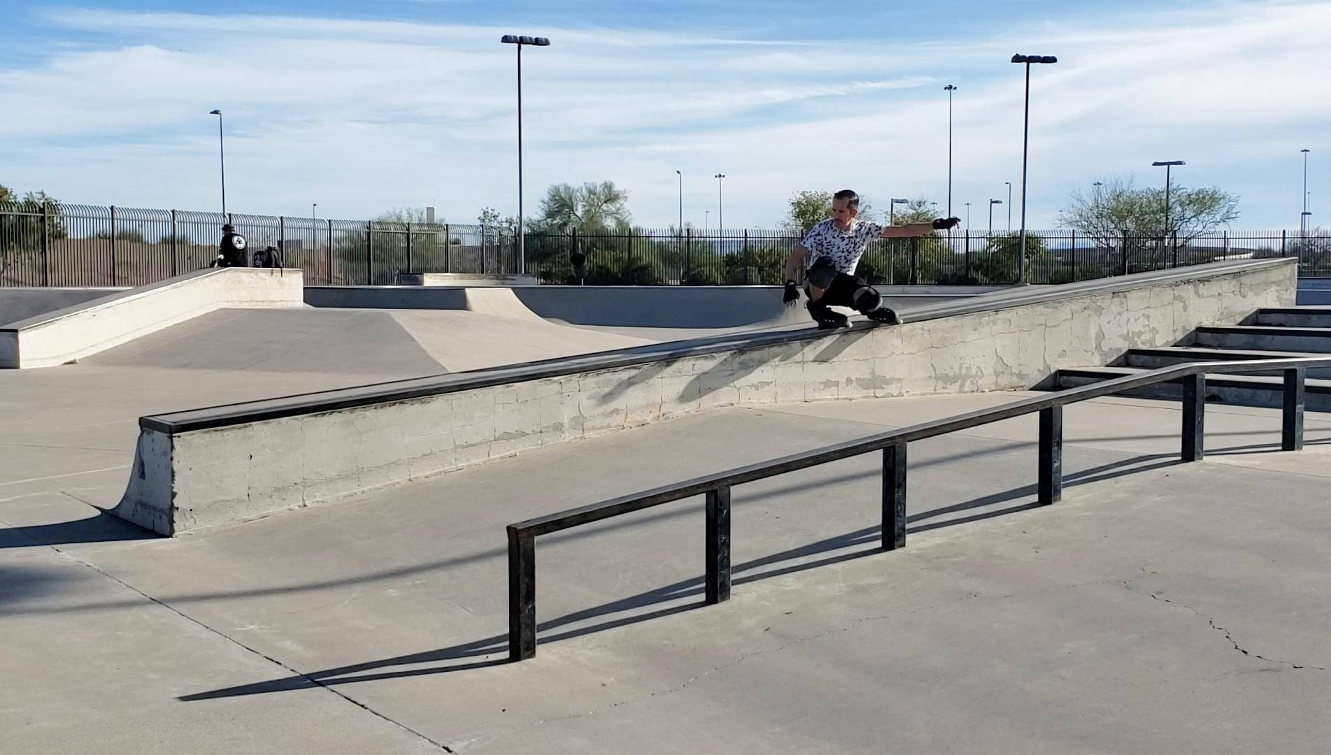 skating 桌布 壁紙 — 一名滑板手在陽光明媚的混凝土滑板公園中，於金屬欄杆上磨滑，背景是清澈的藍天和遠處的樹木。