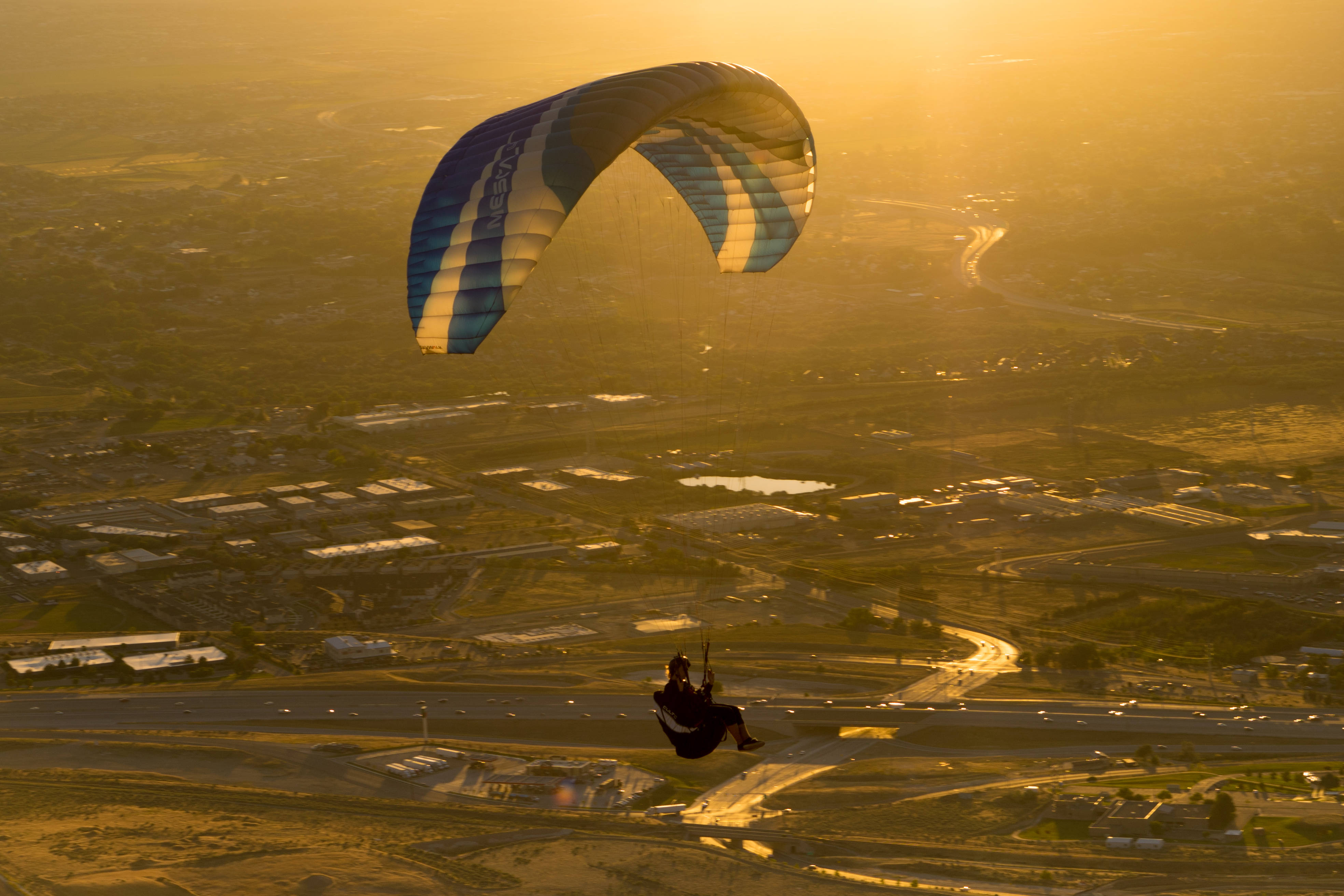 Paragliding 桌布 壁紙 — 一名滑翔傘運動員帶著藍白相間的傘翼，在沐浴著金色夕陽光芒的廣闊城市景觀上空翱翔。