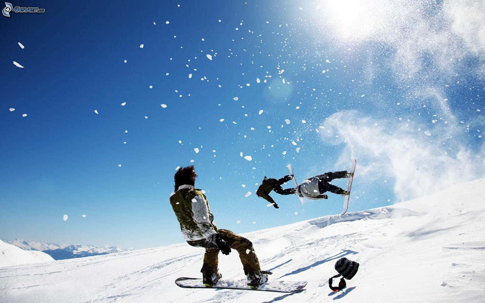 Winter Sports 桌布 壁紙 — 一名滑雪板運動員在半空中表演技巧，另一名滑雪板運動員在陽光燦爛的雪山斜坡上觀看。