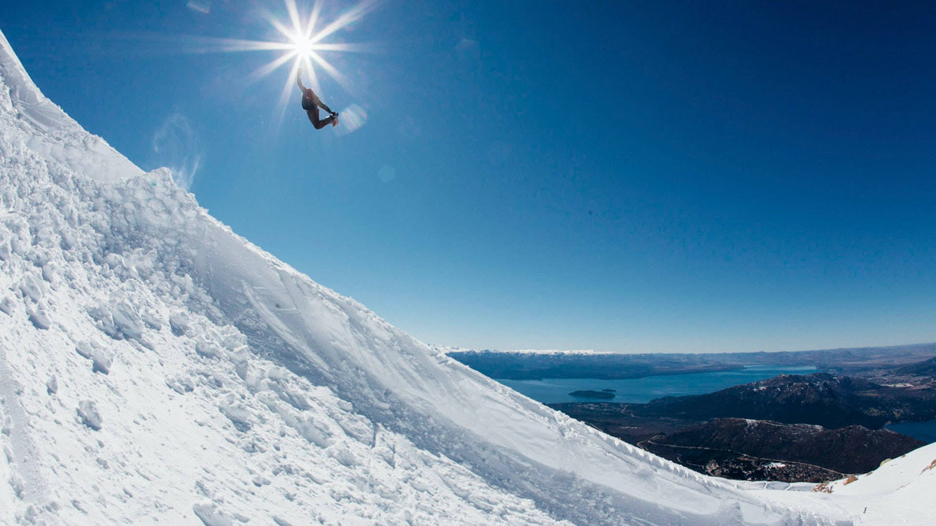 Snowboarding 桌布 壁紙 — 一名滑雪板運動員在清新空氣中翱翔，下方是陽光照耀的雪坡、廣闊的藍色湖泊和遠處的山脈。