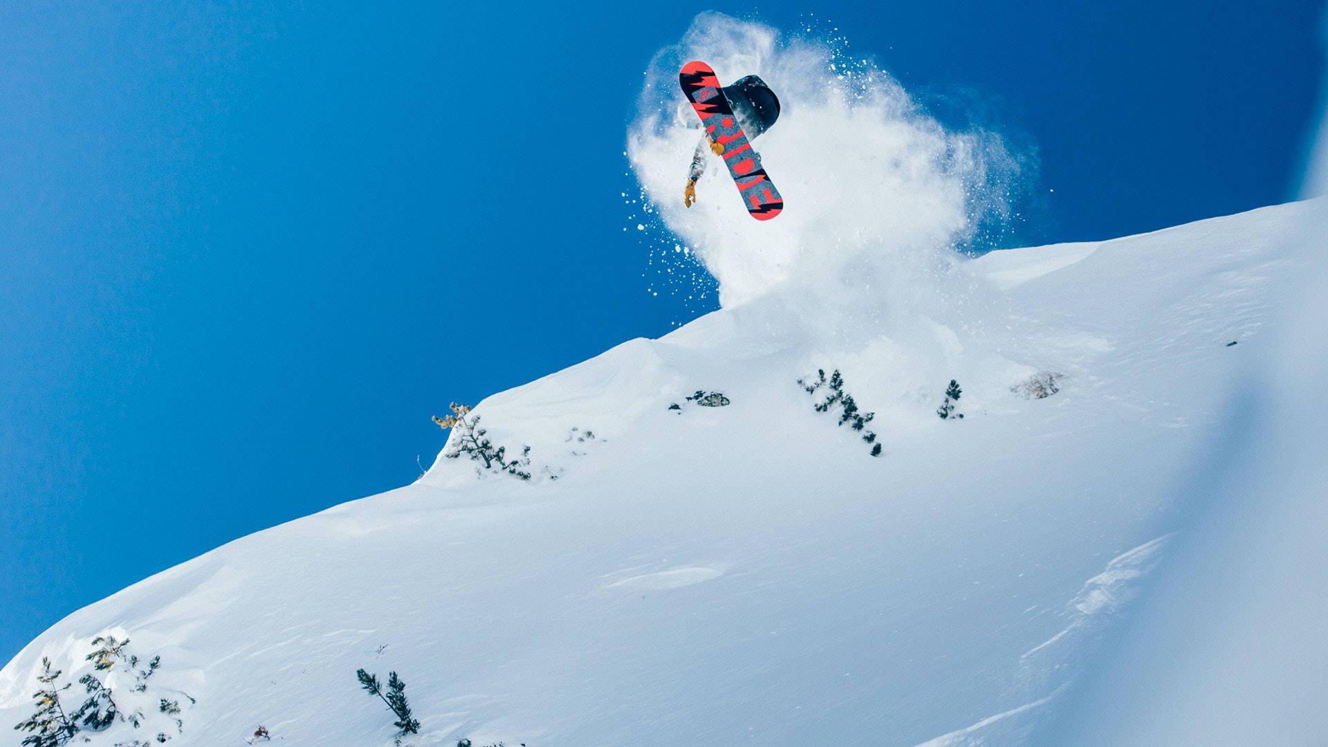 Snowboarding 桌布 壁紙 — 一名滑雪板運動員在清澈的藍天中飛翔，越過白雪覆蓋的山坡，揚起一陣粉雪。