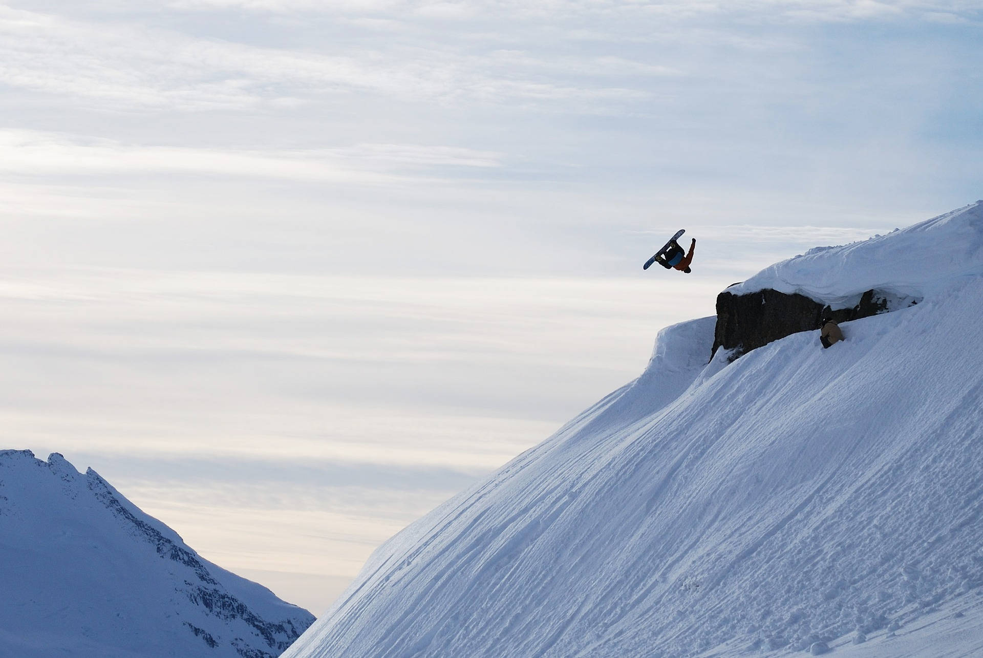Snowboarding 桌布 壁紙 — 一名滑雪板運動員在蒼白的天空下，以廣闊的山脈為背景，從雪崖上做出大膽的後空翻。