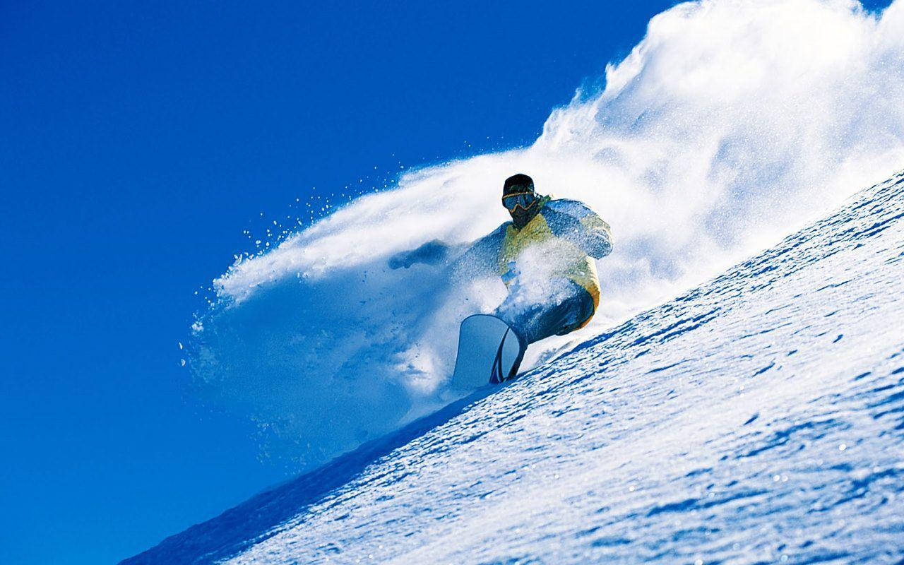 Winter 桌布 壁紙 — 一名滑雪板運動員在陡峭的雪坡上 carve 而下，將大量粉雪噴向清澈的藍天。