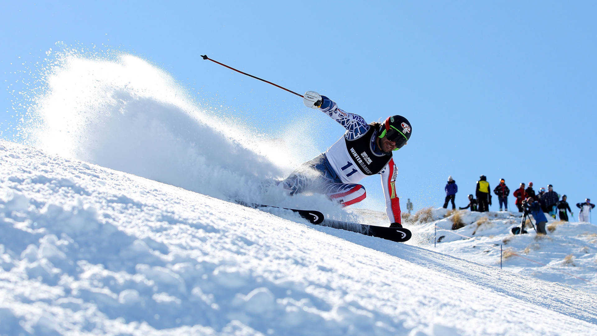 Winter Olympics 桌布 壁紙 — 一名滑雪者急轉彎滑下雪坡，將戲劇性的粉雪噴向明亮的藍天。