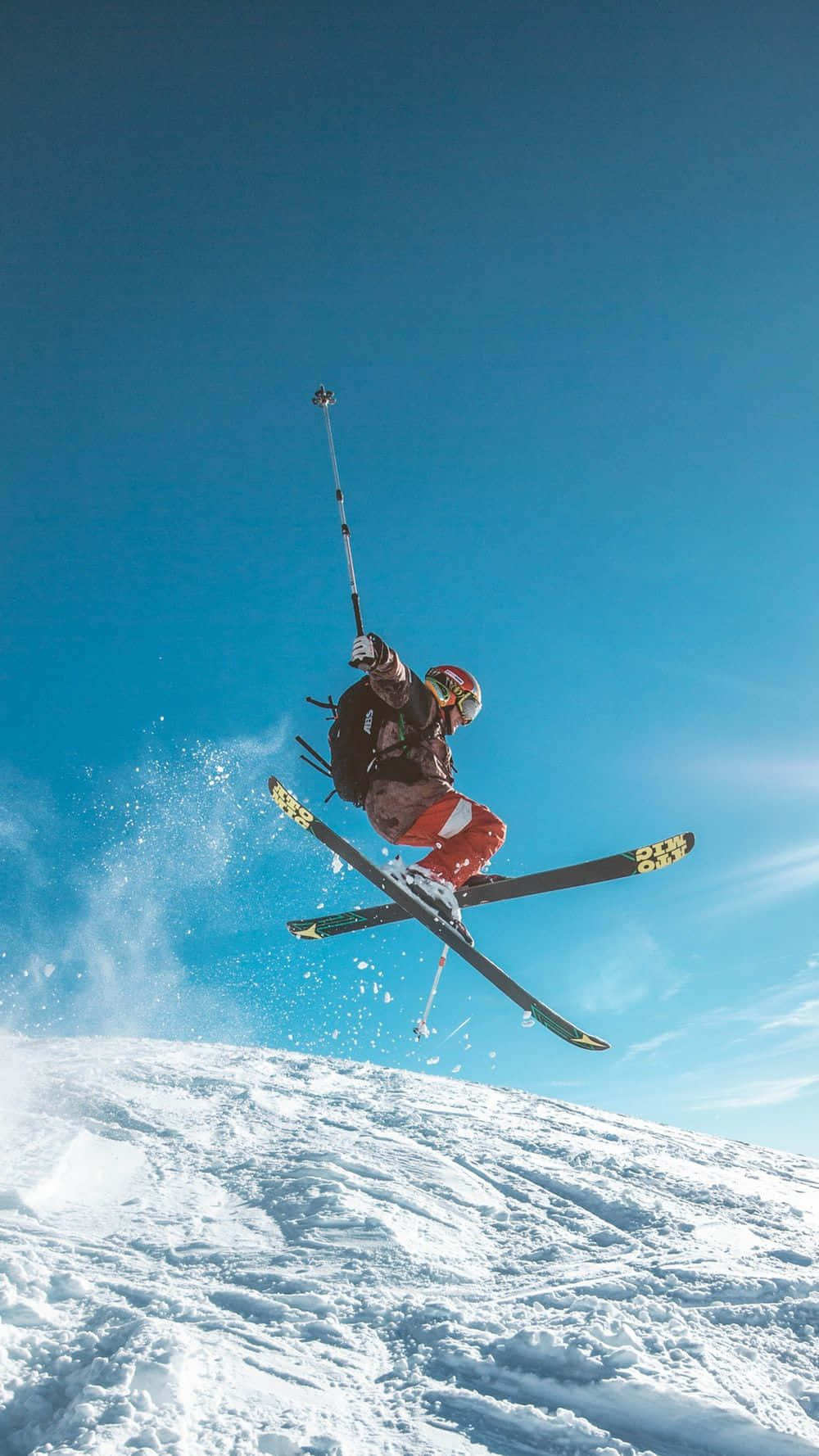 Winter Sports 桌布 壁紙 — 一名滑雪者飛越清澈藍天，在積雪的山坡上空翱翔，滑雪板濺起雪花。