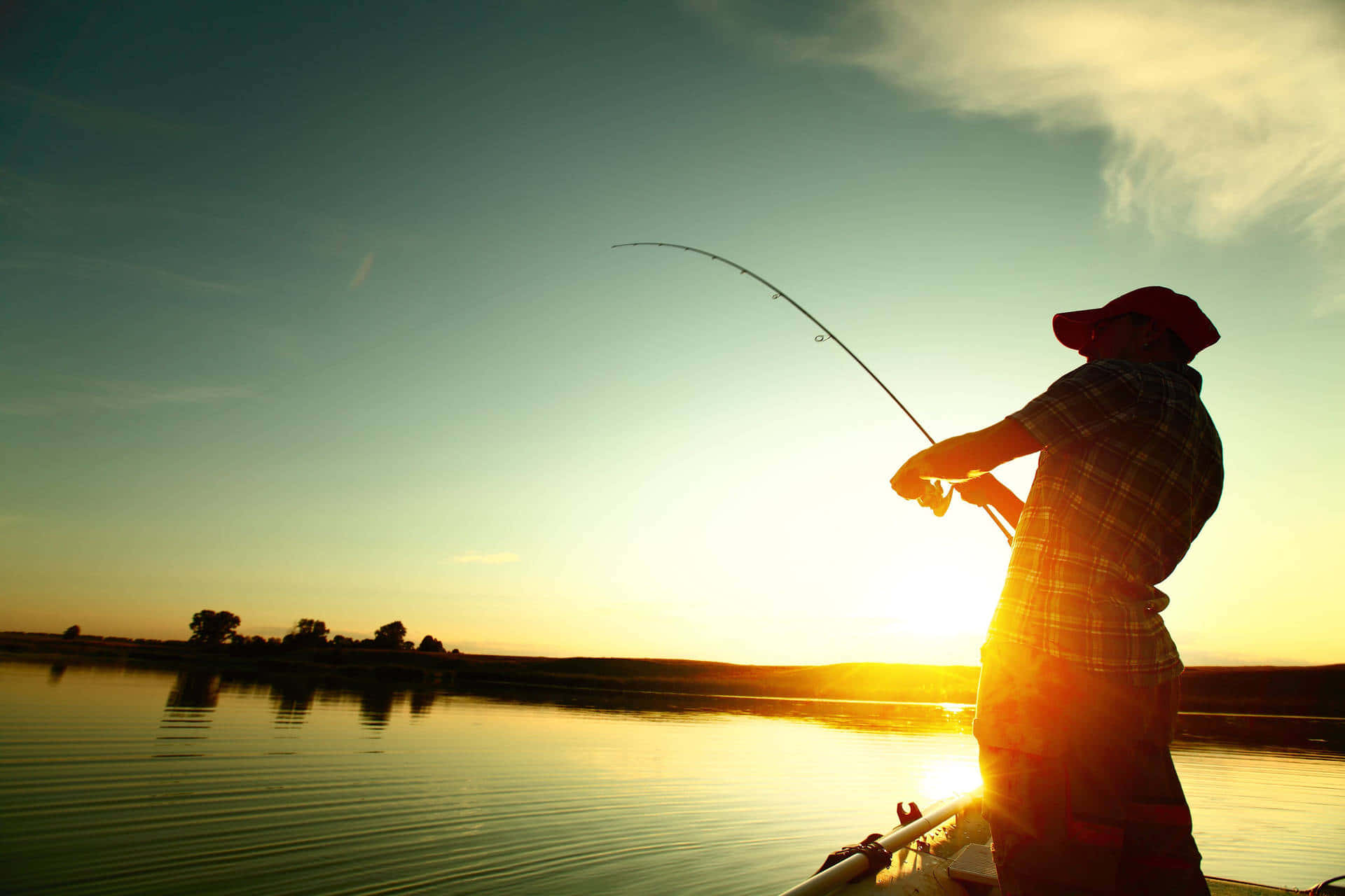 Fishing Rod 桌布 壁紙 — 一名漁夫在日落時分從船上拋出釣線，金色光芒映照在平靜的水面上，遠方樹木呈現剪影。