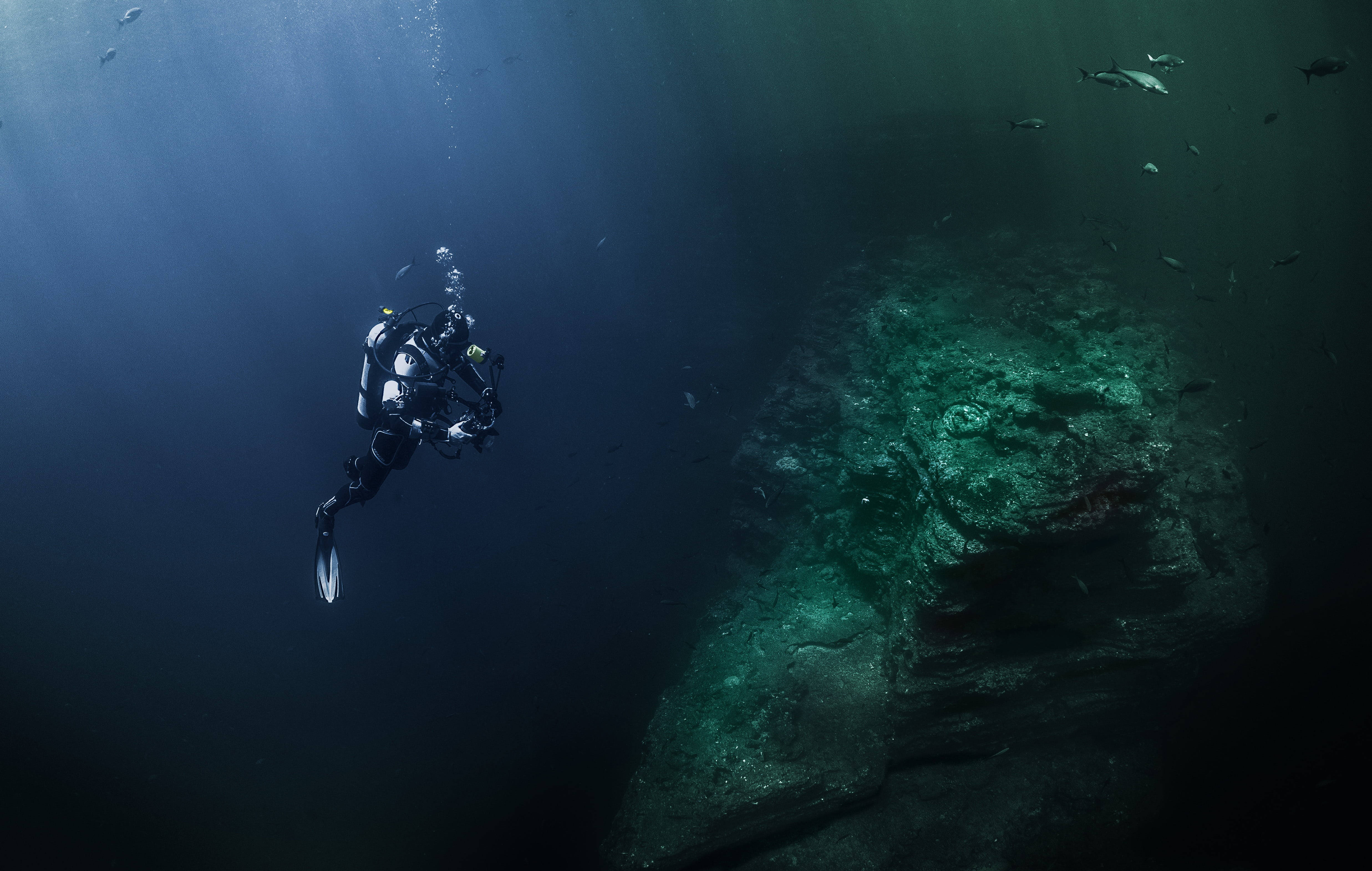 diving 桌布 壁紙 — 一名潛水員探索陽光照射的水下遺跡，魚群在深藍綠色的水中游近。