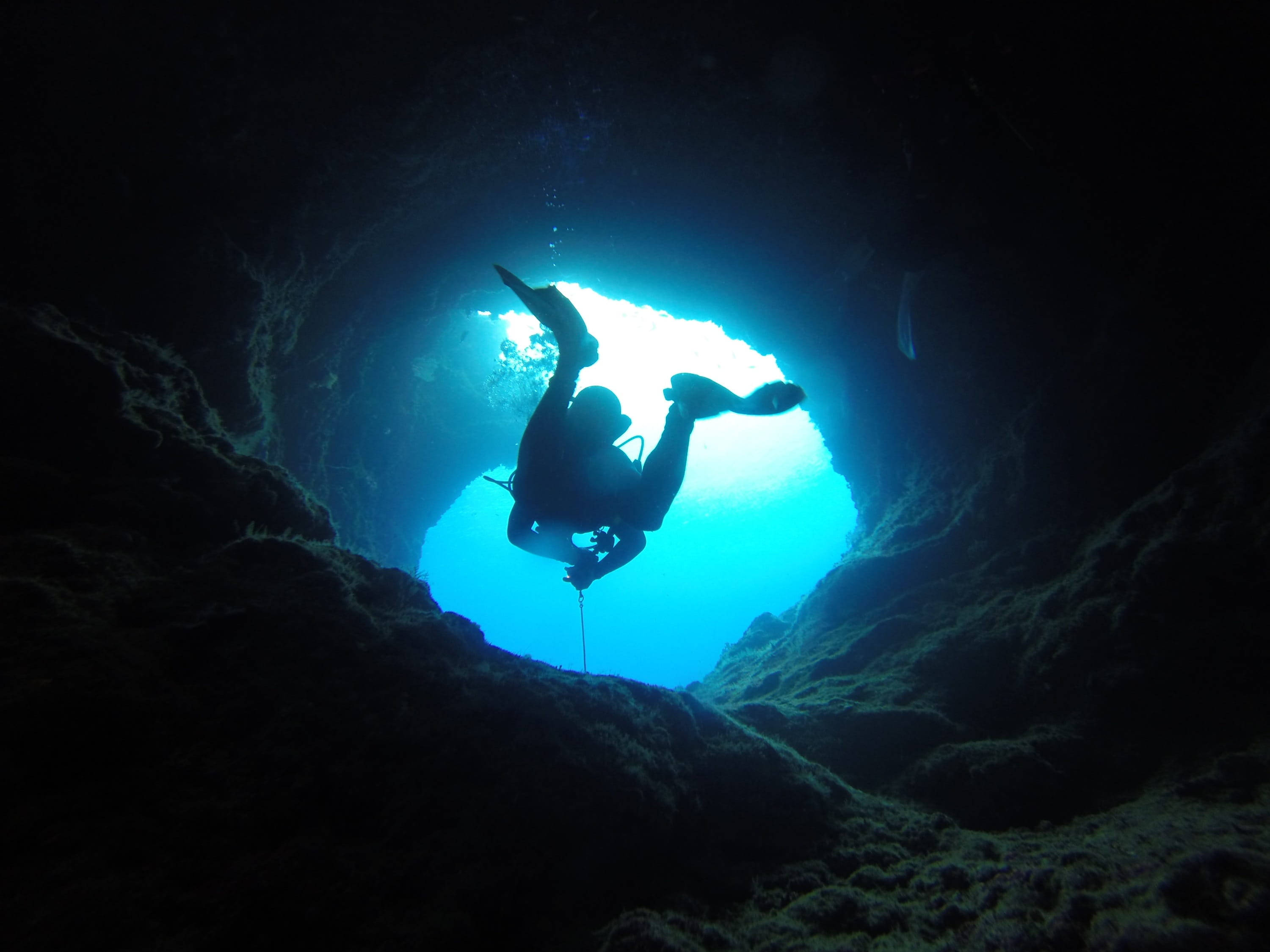 diving 桌布 壁紙 — 一名潛水員的剪影在探索黑暗的水下洞穴，明亮的藍光從上方的開口湧入。