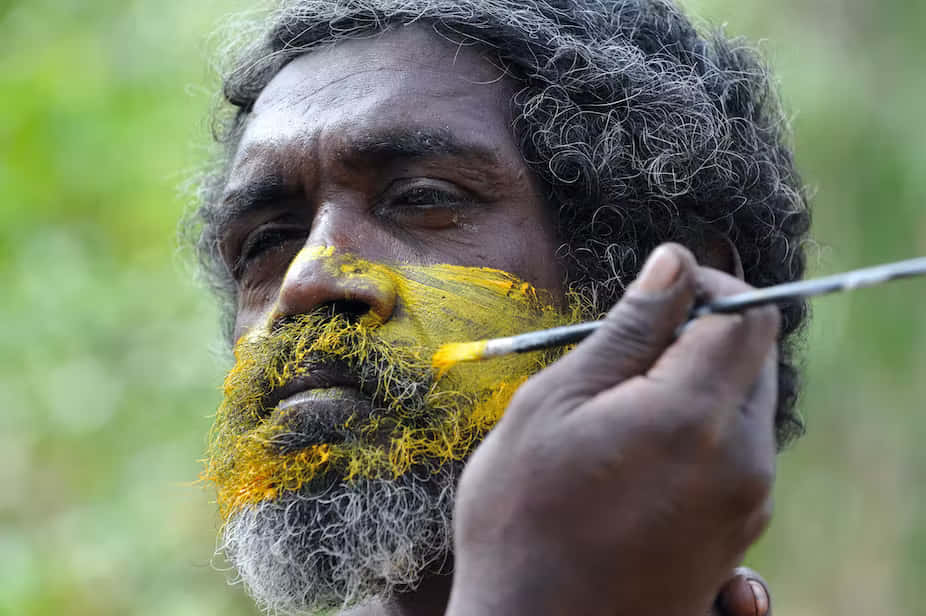 Aboriginal 桌布 壁紙 — 一名灰色捲髮和鬍鬚的男子，在模糊的綠色背景下，用刷子將鮮黃色的顏料塗在臉上。