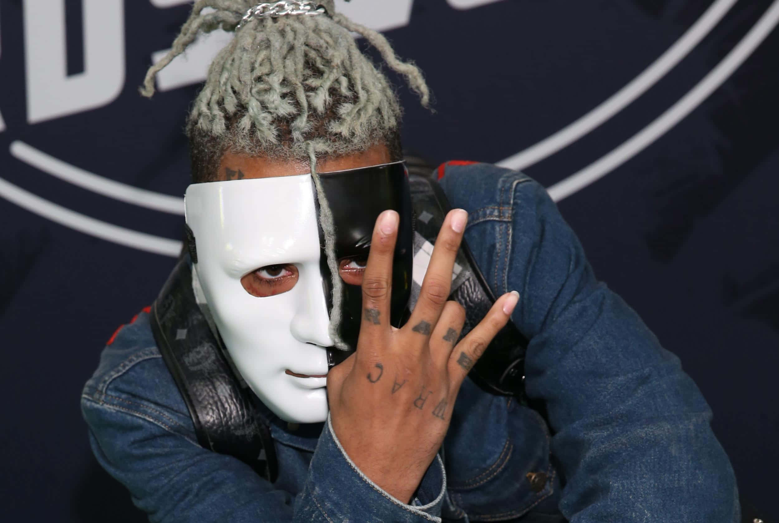 XXXTENTACION 桌布 壁紙 — 一名灰色辮子頭的人戴著黑白分開的面具，用有刺青的手拿著它，背景是深藍色的品牌背景。