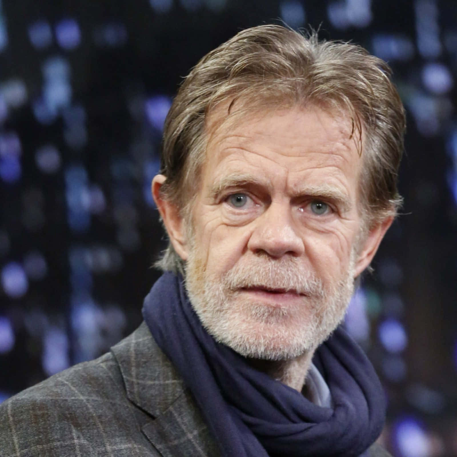 william h. macy 桌布 壁紙 — 一名灰髮灰鬚男子戴著深色圍巾，以模糊的藍色散景背景為襯托的特寫肖像。