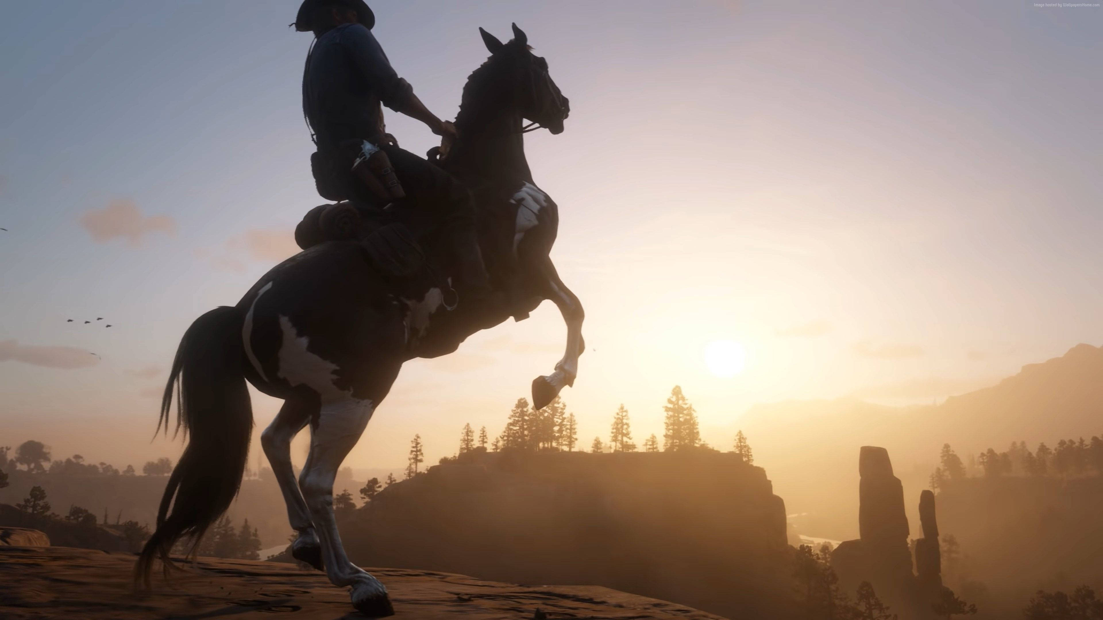 Red Dead Redemption 2 Desktop 桌布 壁紙 — 一名牛仔在日落時分騎著一匹黑白相間的馬，背景是金色的天空和遠山。
