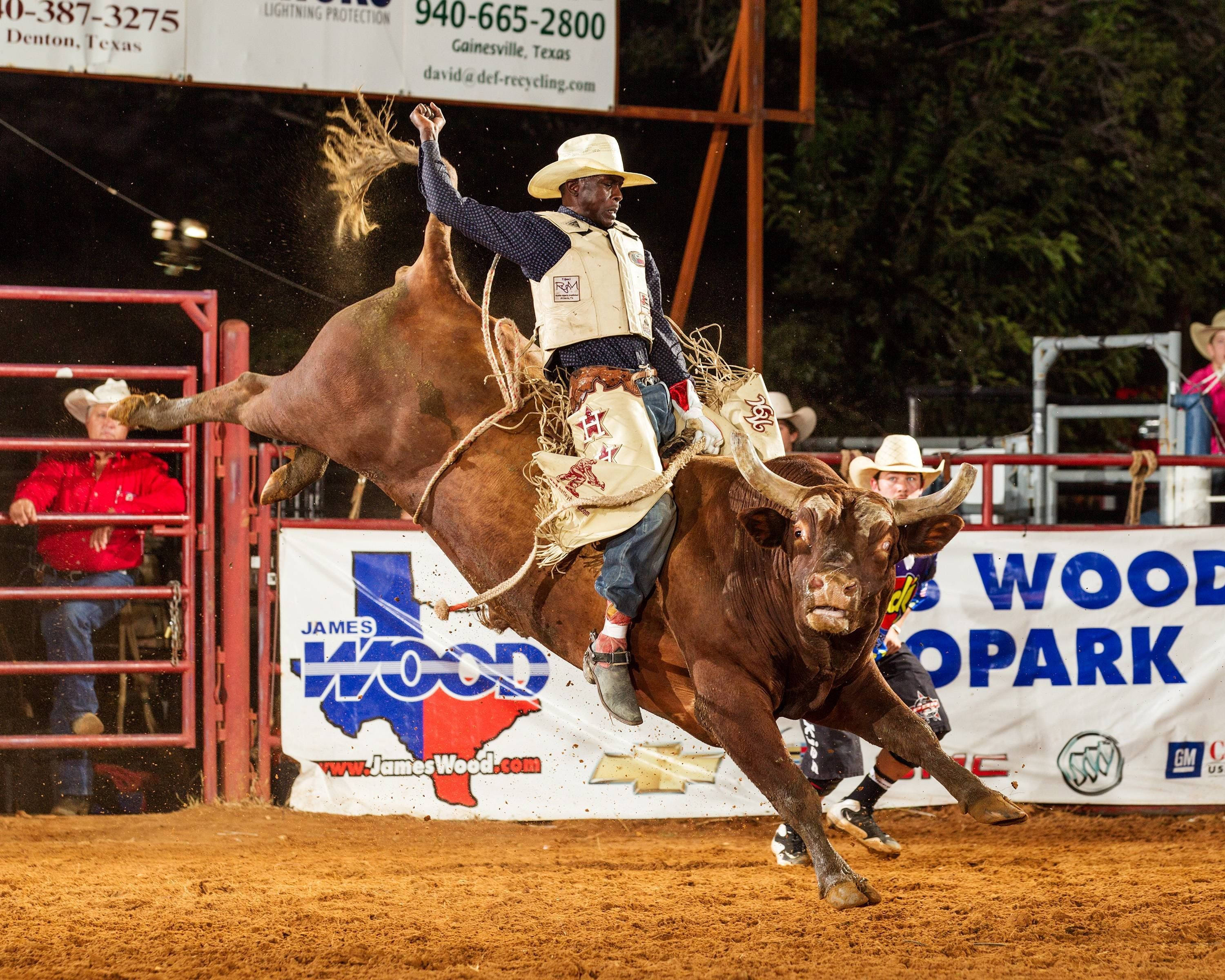 Bull Riding 桌布 壁紙 — 一名牛仔在體育場燈光下，在設有觀眾和橫幅的牛仔競技場中緊抓著一頭狂跳的公牛。