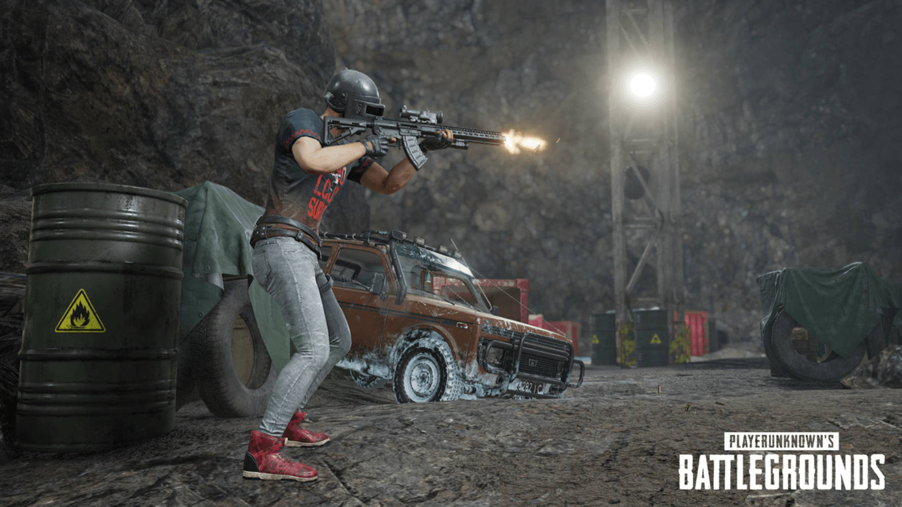 絕地求生 桌布 壁紙 — 一名玩家在《PUBG》中，於光線昏暗的洞穴內，從一輛生鏽的汽車後方開槍射擊，現場有工業用桶和明亮的頭頂燈光。