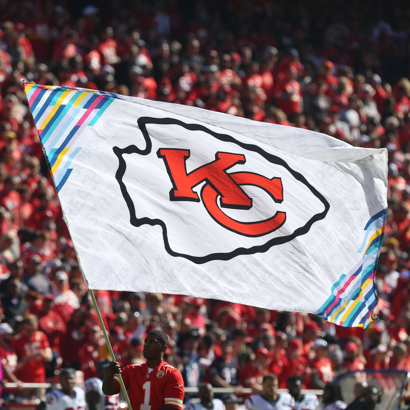 Kansas City Chiefs Logo 桌布 壁紙 — 一名球迷在陽光明媚的比賽中，於座無虛席的體育場內揮舞一面大型白色堪薩斯城酋長隊旗幟，旗上有紅色標誌和彩色條紋。