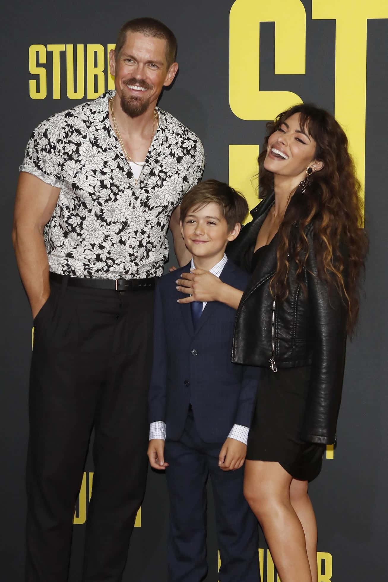 sarah shahi 桌布 壁紙 — 一名男子、女子和小孩在電影首映會上，在搶眼的黃色背景前一起微笑。