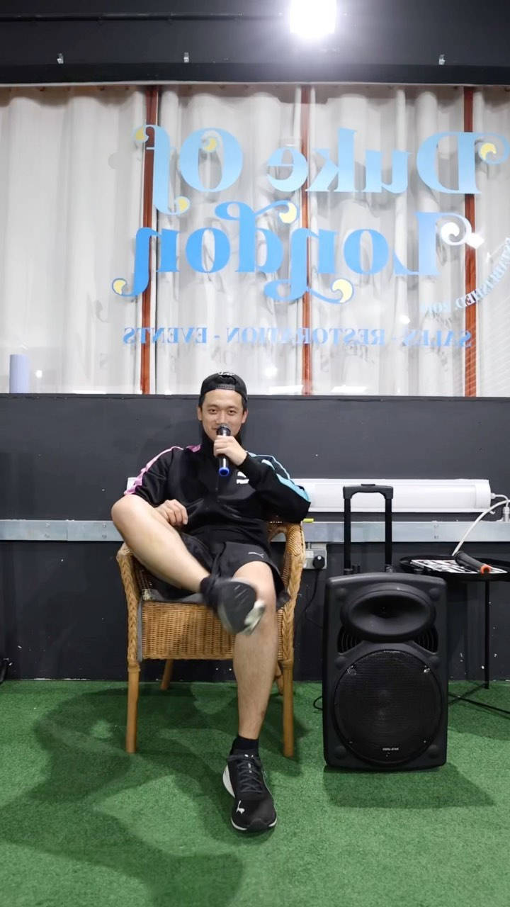 人物 桌布 壁紙 — 一名男子休閒地坐在藤椅上，在明亮的舞台燈光下，於人造草地上拿著麥克風，旁邊有一個大型揚聲器。