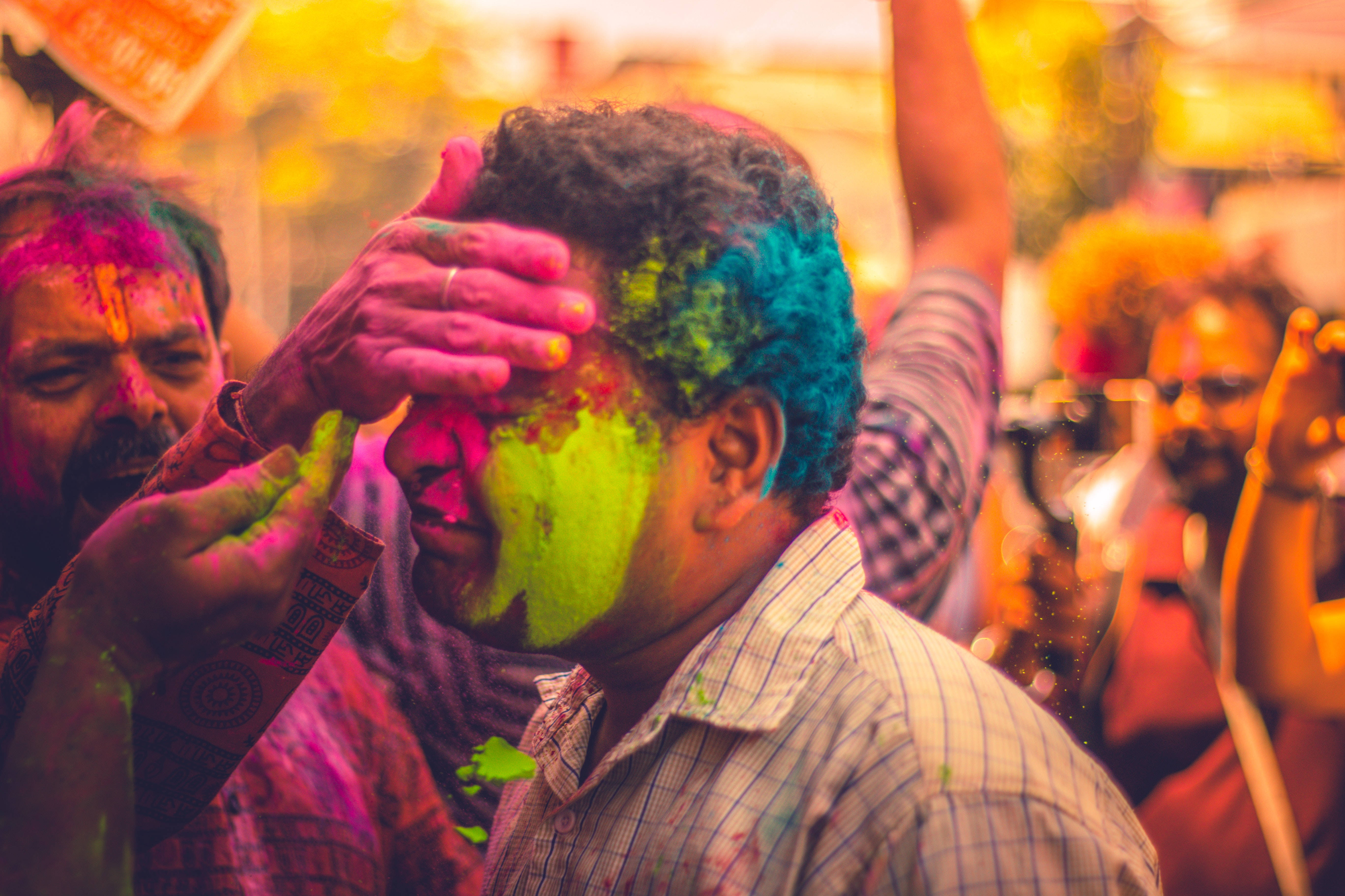 Holi 桌布 壁紙 — 一名男子全身沾滿鮮豔的彩色粉末，臉上塗有綠色和粉紅色的粉末，周圍的人們正在慶祝。