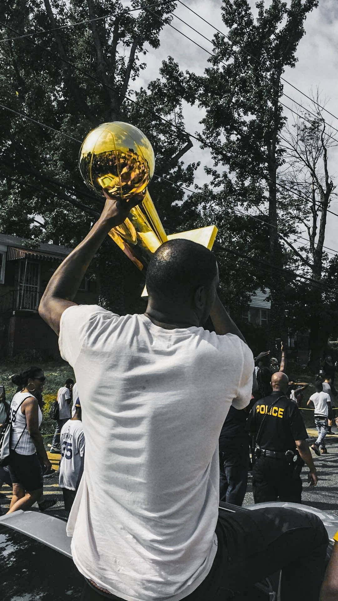 Trophy 桌布 壁紙 — 一名男子勝利地高舉閃閃發光的金色獎盃，坐在一輛汽車上，身處陽光明媚的街道，周圍是歡呼的人群和警察。